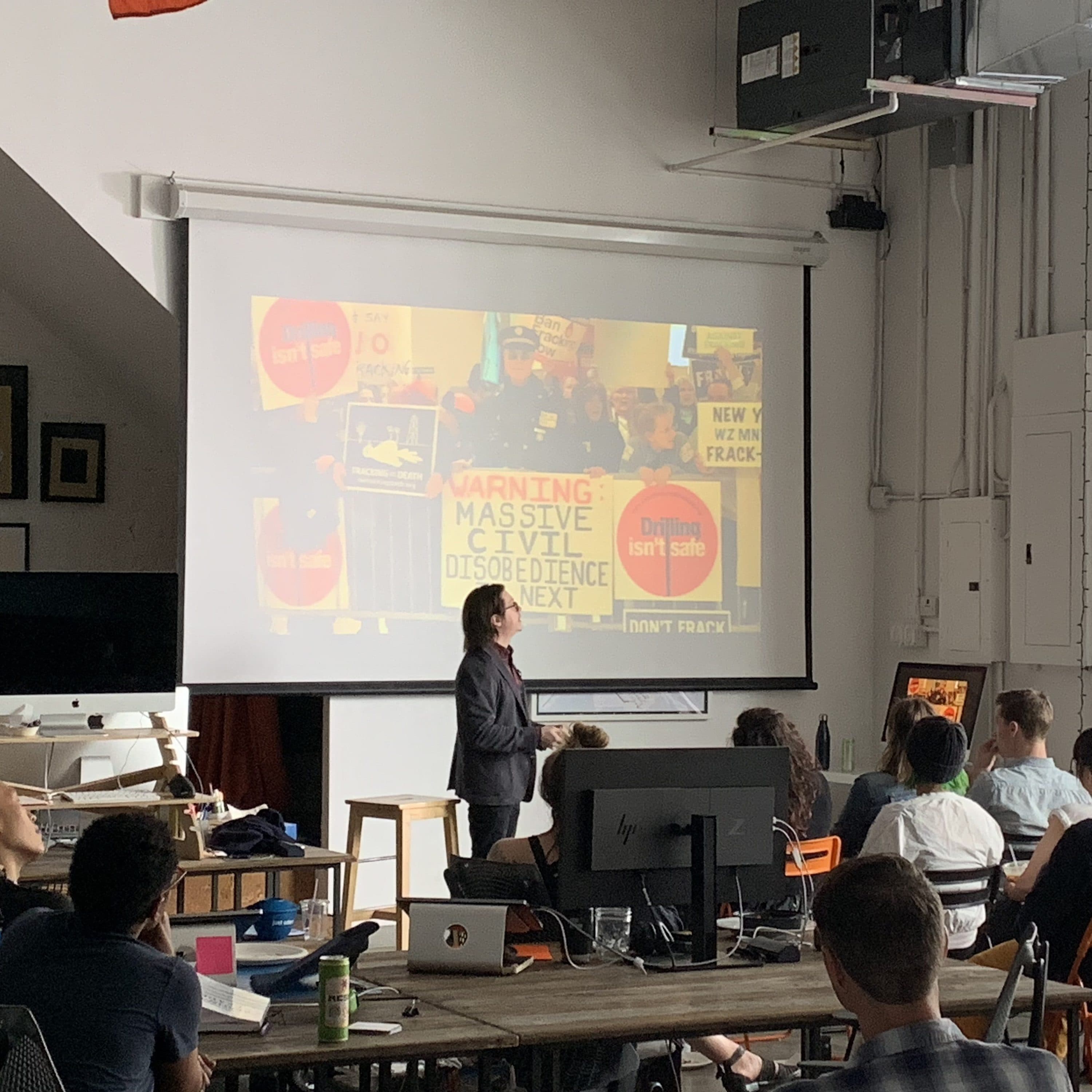 A person is presenting to an audience in a classroom setting. The presentation slide displays protest signs with messages like "Warning: Massive Civil Disobedience Next" and "Earth Isn't Safe." Various objects and artworks adorn the room.