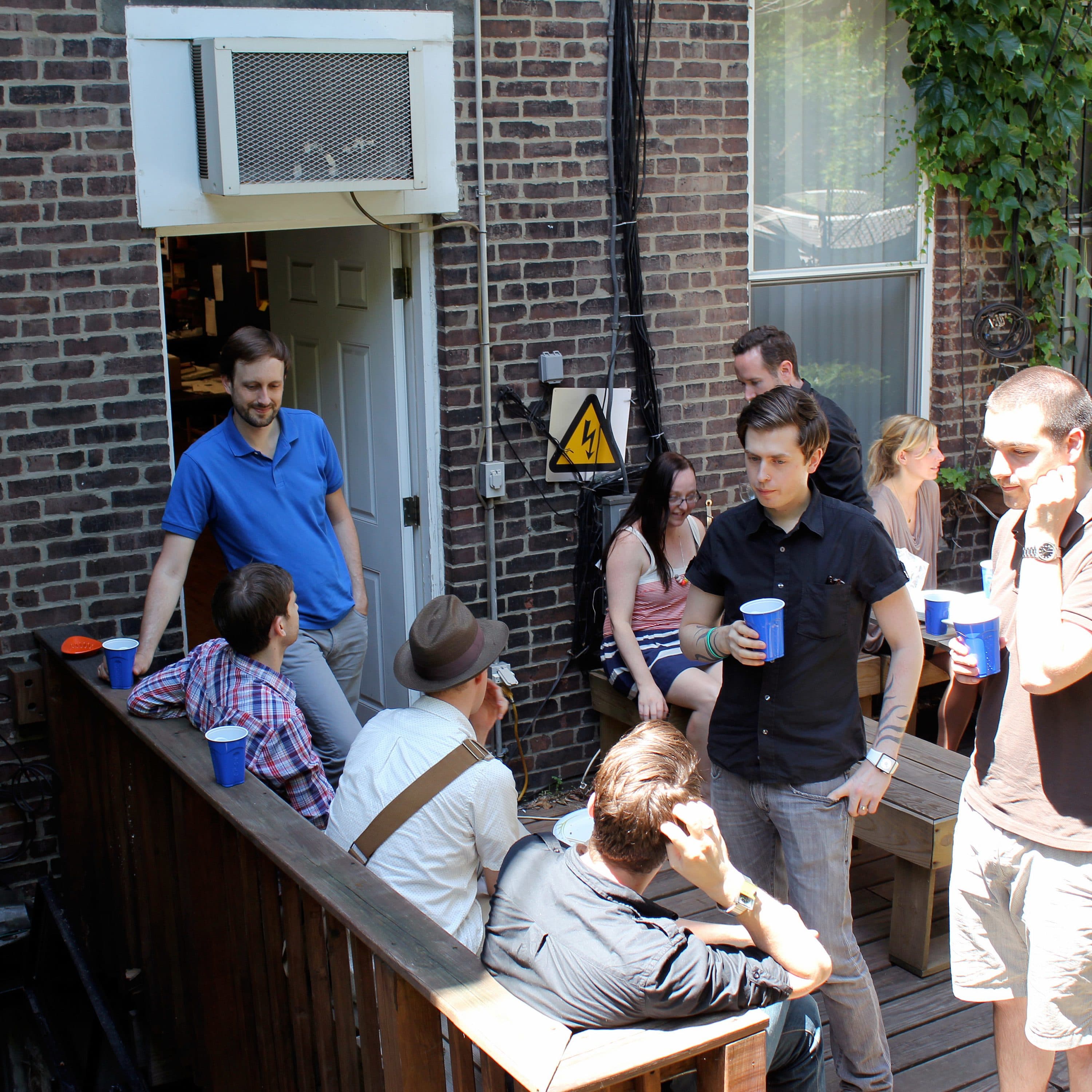A group of people is gathered on a wooden deck outside a brick building. Some are seated, and others are standing, holding blue cups. The scene appears casual and social. One person is entering or exiting through a door in the background. Sunlight illuminates the area.