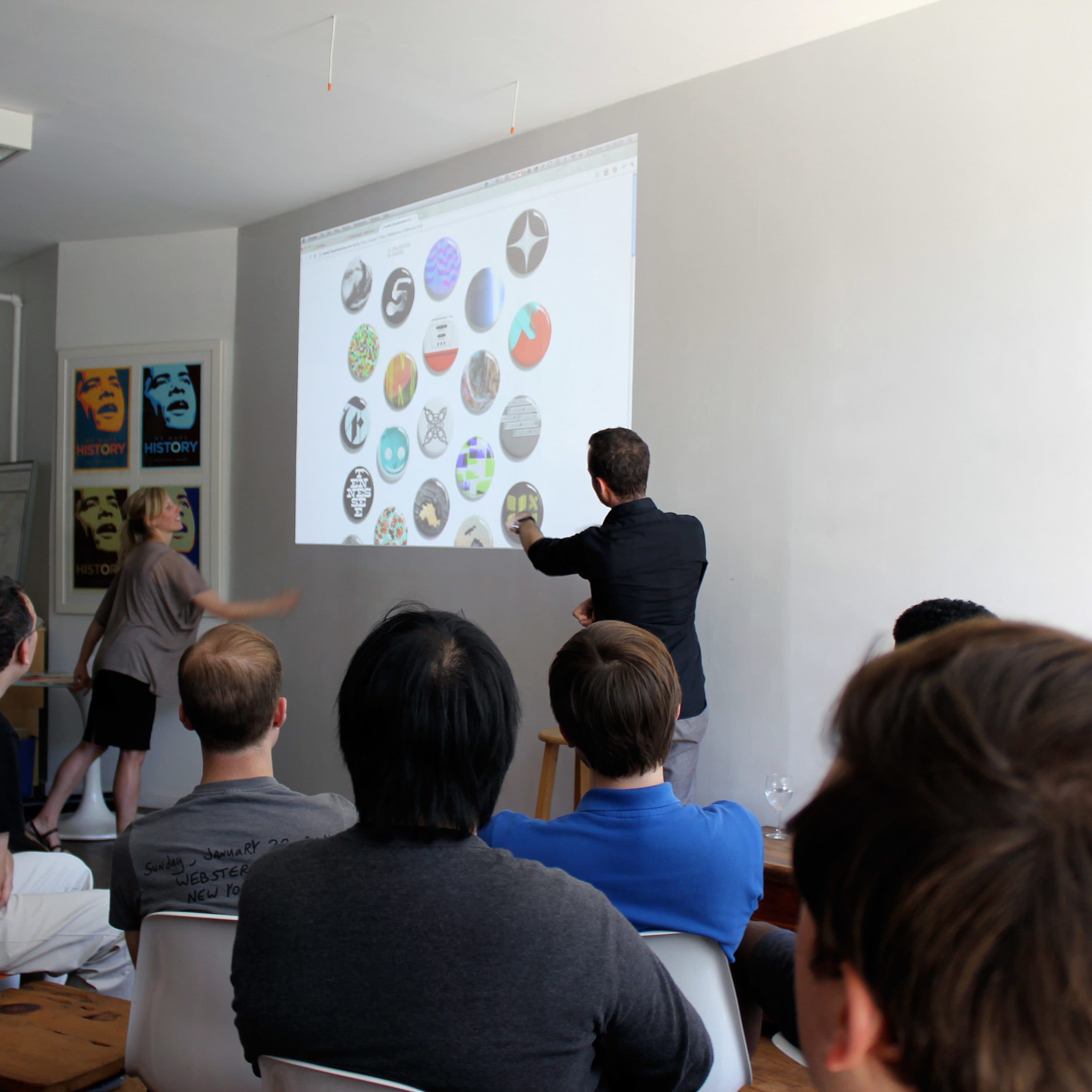 A group of people is seated in a room, attentively watching two individuals at the front who are pointing at a projected screen displaying various circular logos. The room has white walls, posters on the left, and a mix of seating, including chairs and a couch.