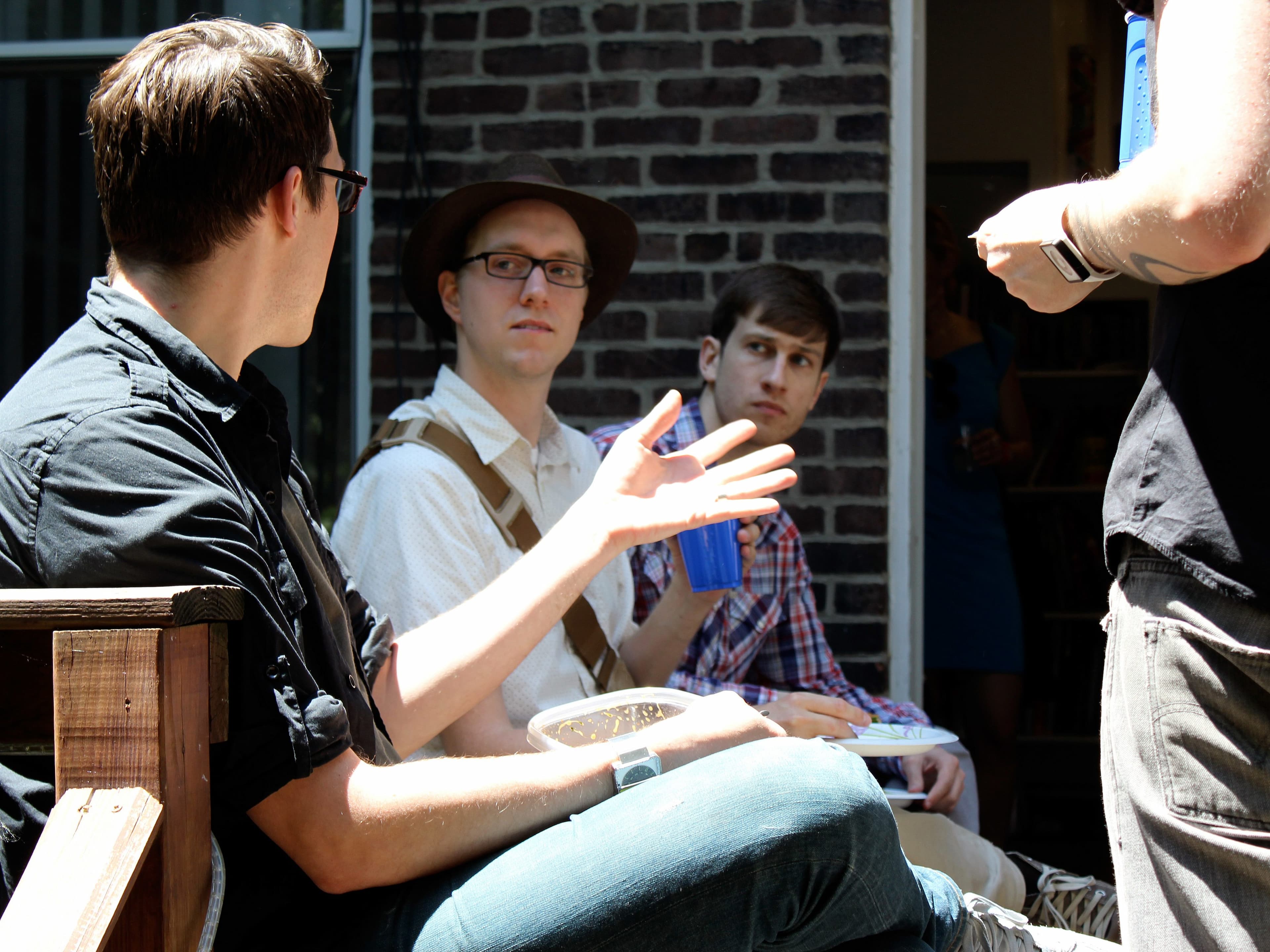 Three people are seated outdoors, engaged in a conversation, with one gesturing animatedly and holding a cup. Another person, standing and facing away from the camera, appears to be joining the discussion. They are in a space with a brick wall and a sunny ambiance.