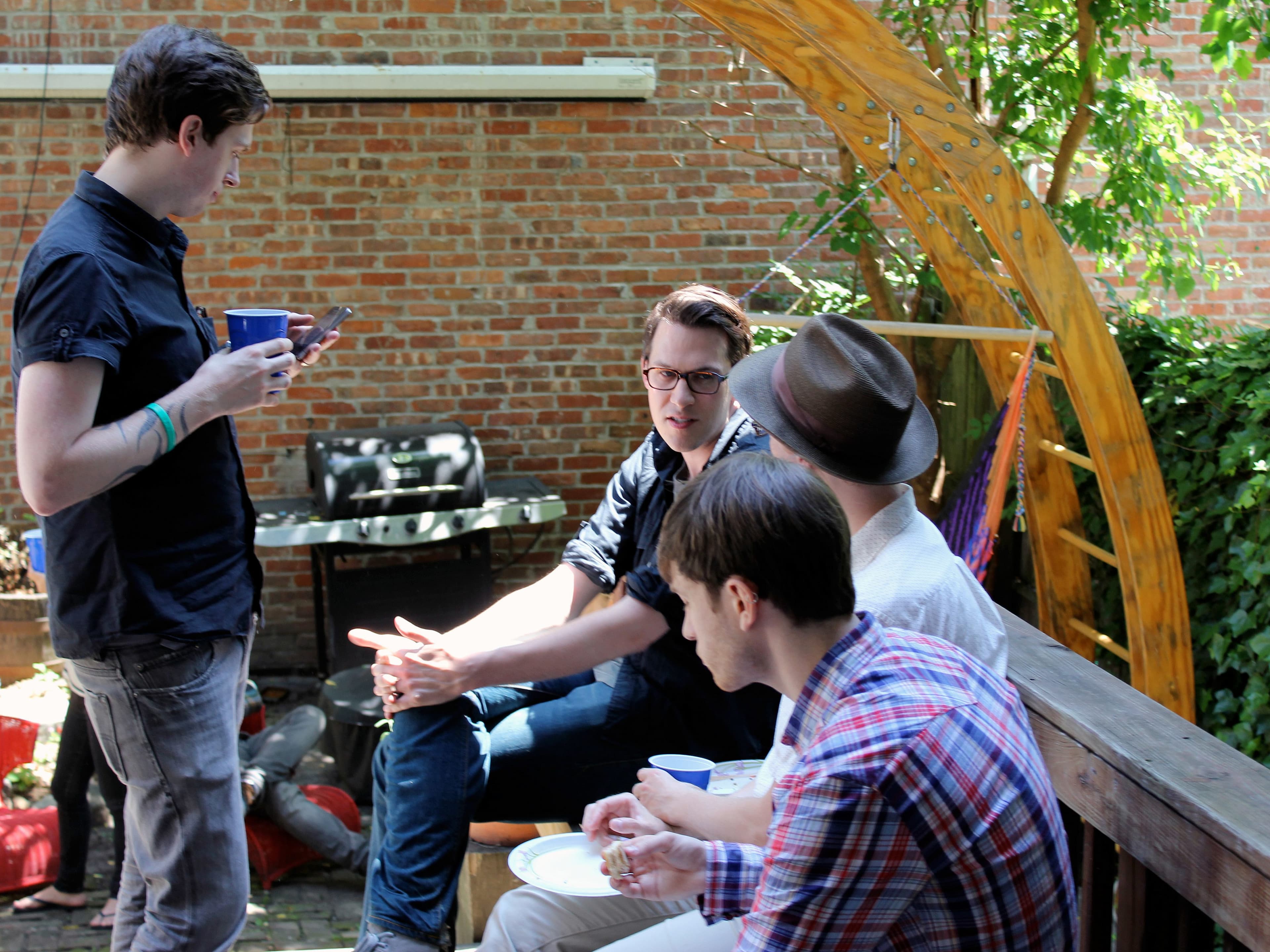 A group of four young adults socializing outdoors. One person is standing and looking at their phone while holding a cup. Three others are seated on a wooden bench, involved in conversation. There's a wooden trellis and green foliage in the background.