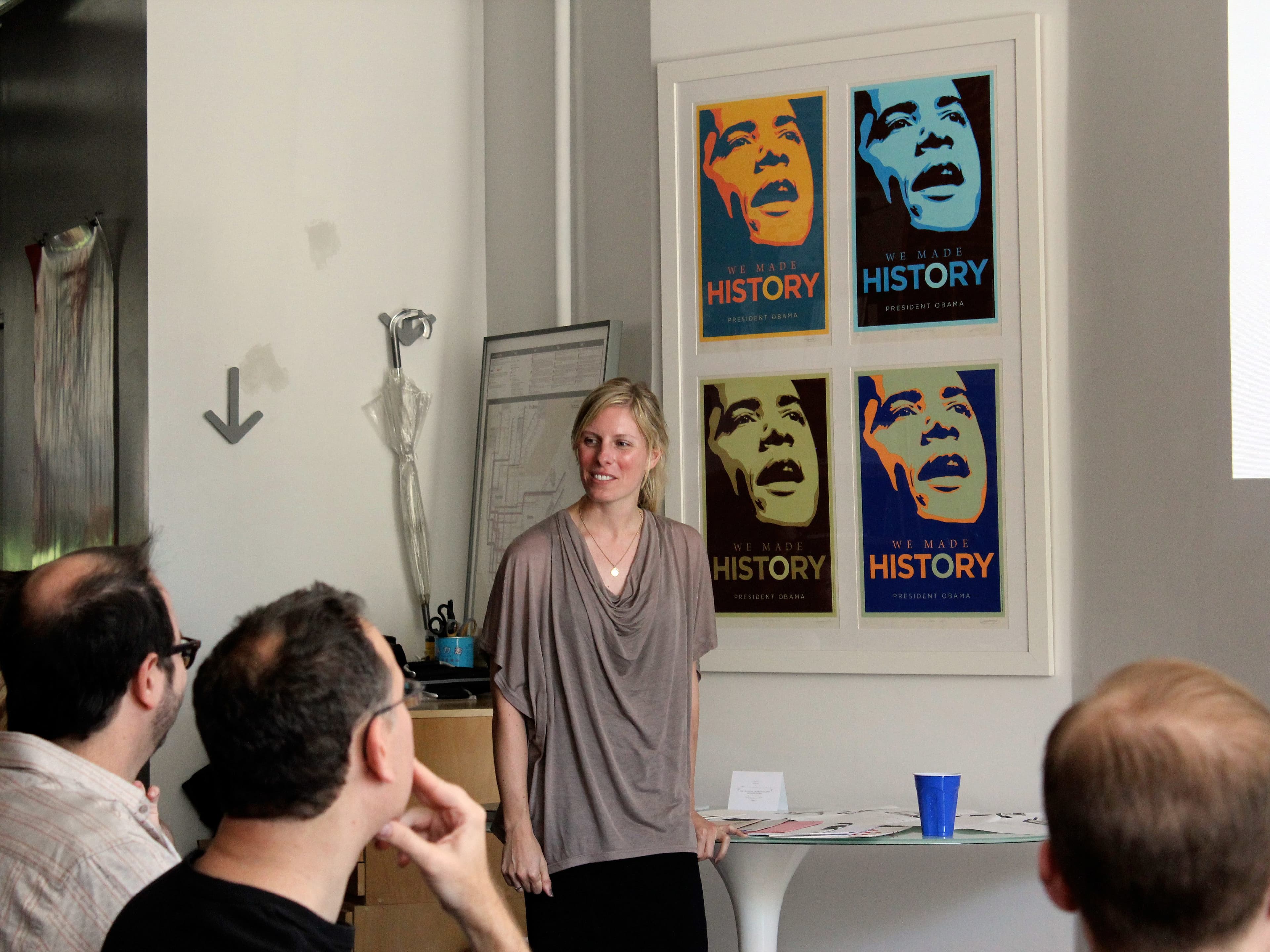 A woman stands in front of a group of seated people, giving a presentation. Behind her on the wall are four framed posters with the phrase "We Made History" and stylized images of a man. A blue cup and some papers are on the table beside her.