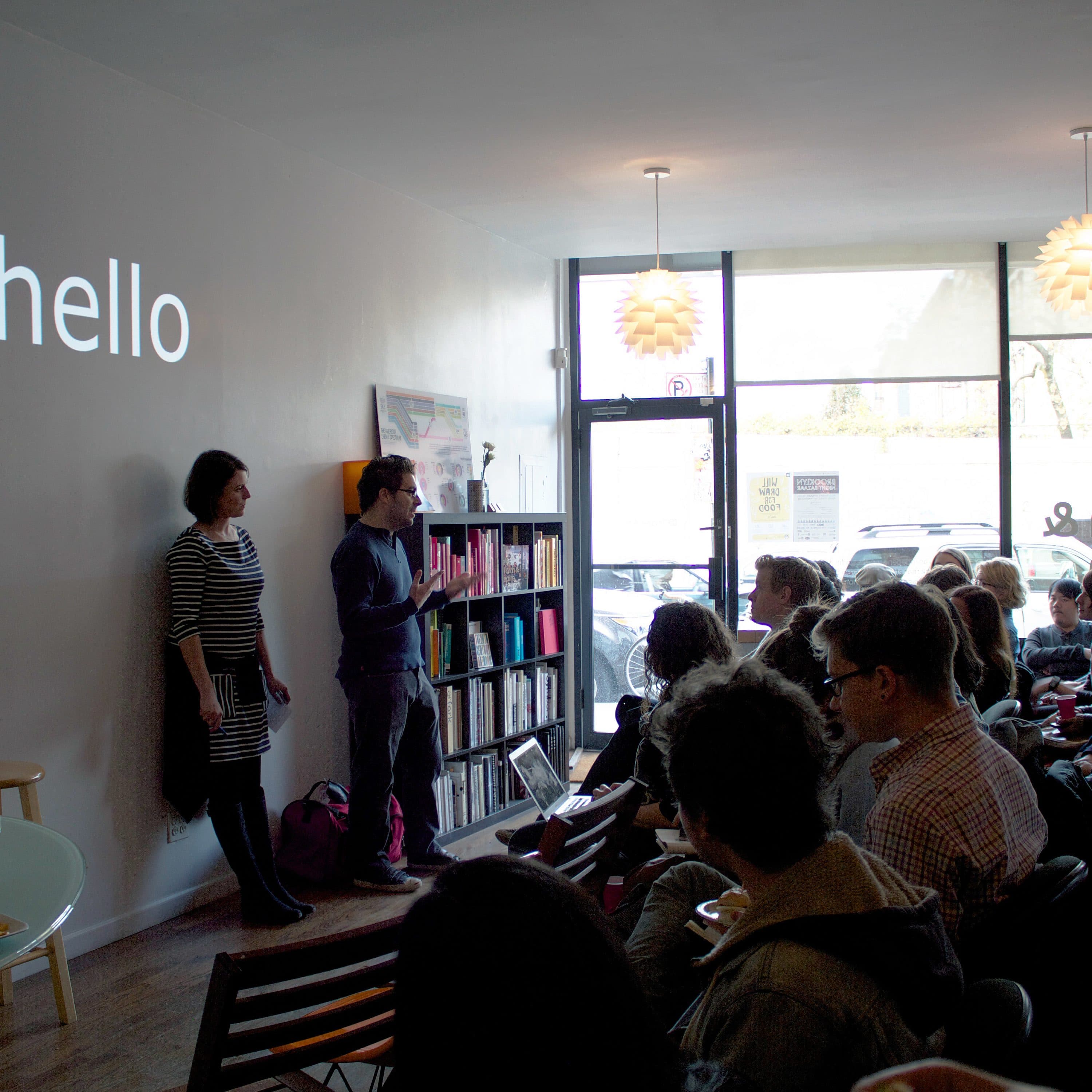 A woman and a man are standing at the front of a small room, facing an audience seated in rows. The word "hello" is projected on the wall behind them. The room has a minimalist design with hanging lamps and large windows allowing natural light.