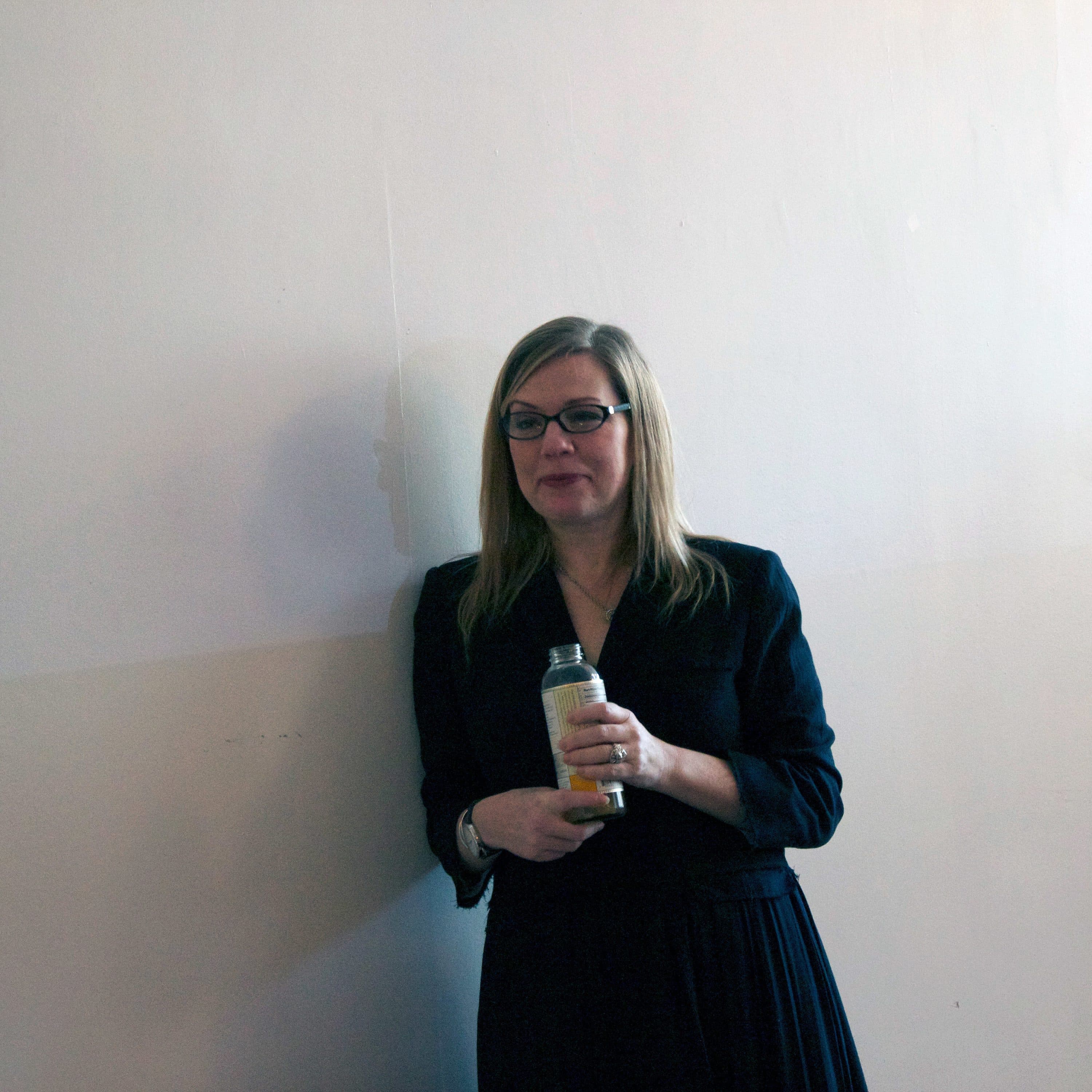 A woman with long blonde hair, glasses, and a black dress stands against a light-colored wall holding a bottled drink. The image is minimally decorated, with some yellow flowers visible in the lower left corner.