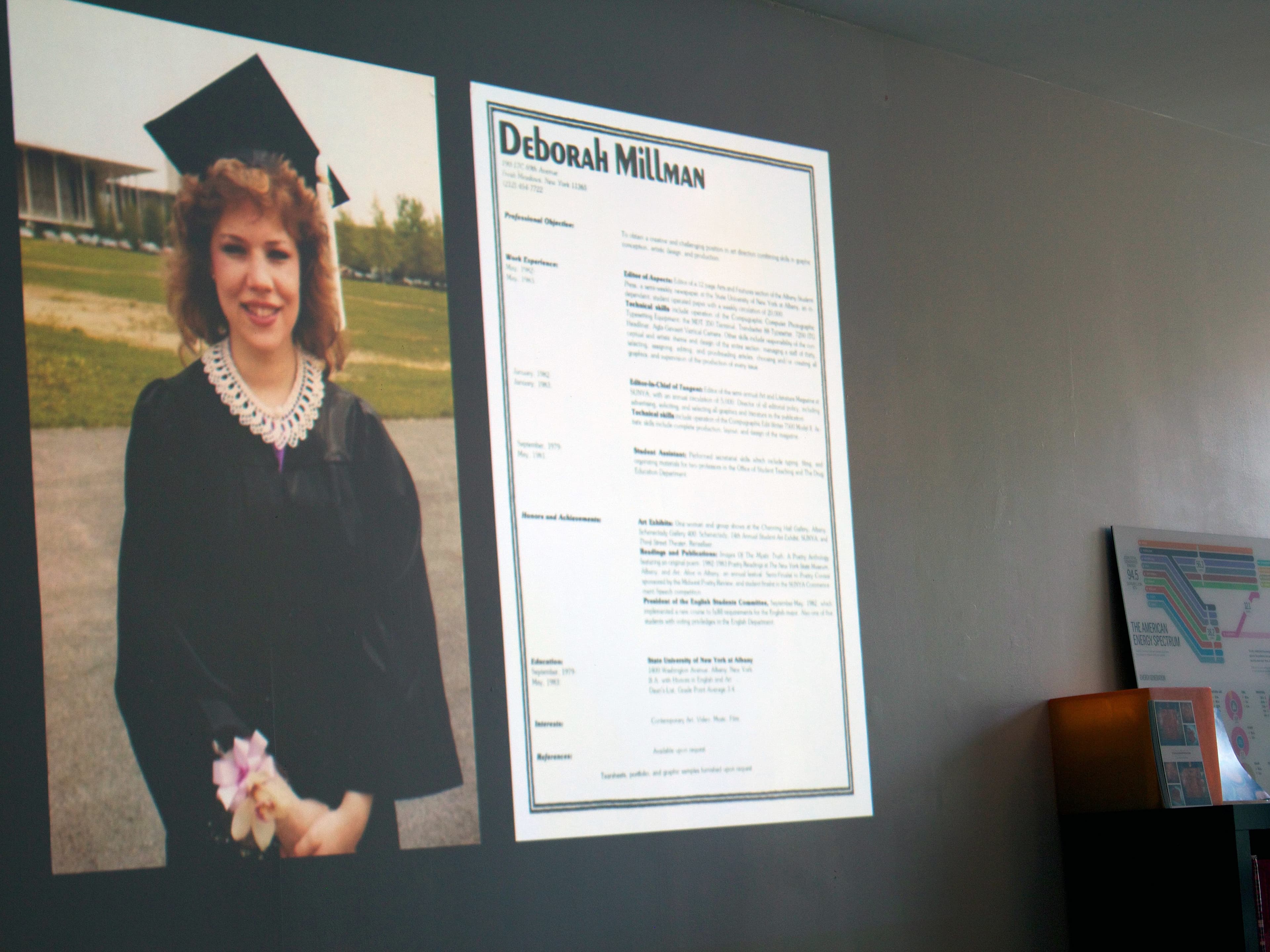 A projection on a wall shows a woman in a graduation cap and gown holding a diploma. Beside her image is a résumé for "Deborah Millman" with details of her education, skills, and experience. In the background is a shelf and a framed piece of artwork.