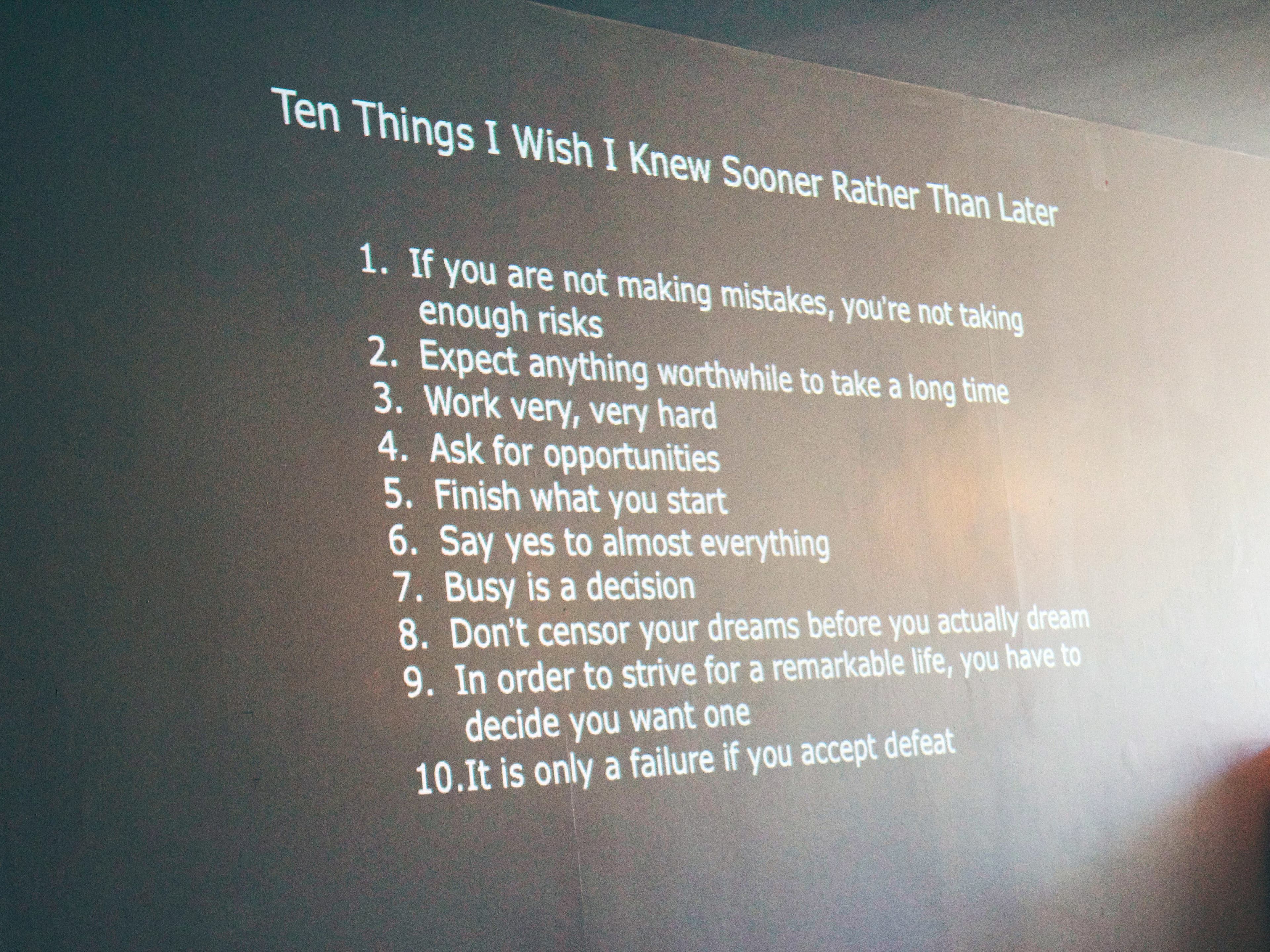 A projected list titled "Ten Things I Wish I Knew Sooner Rather Than Later" on a wall. The list includes advice such as taking risks, working hard, asking for opportunities, finishing what you start, and not censoring dreams. The room is dimly lit.