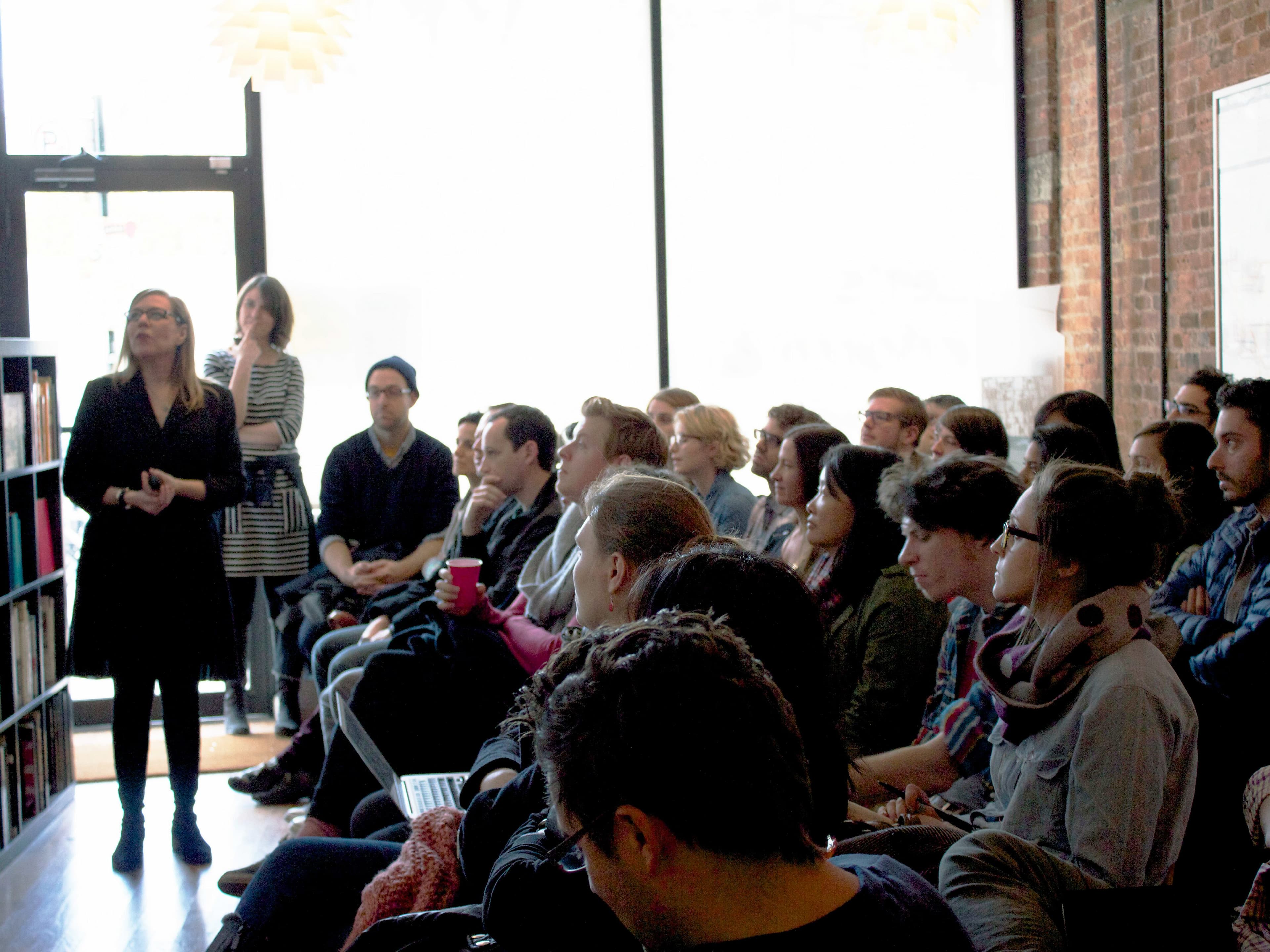 A speaker stands at the front of a room filled with attentive people sitting and listening. The room has large windows letting in natural light, and shelves with books are visible on the left. The audience is diverse, with some people taking notes and others looking engaged.