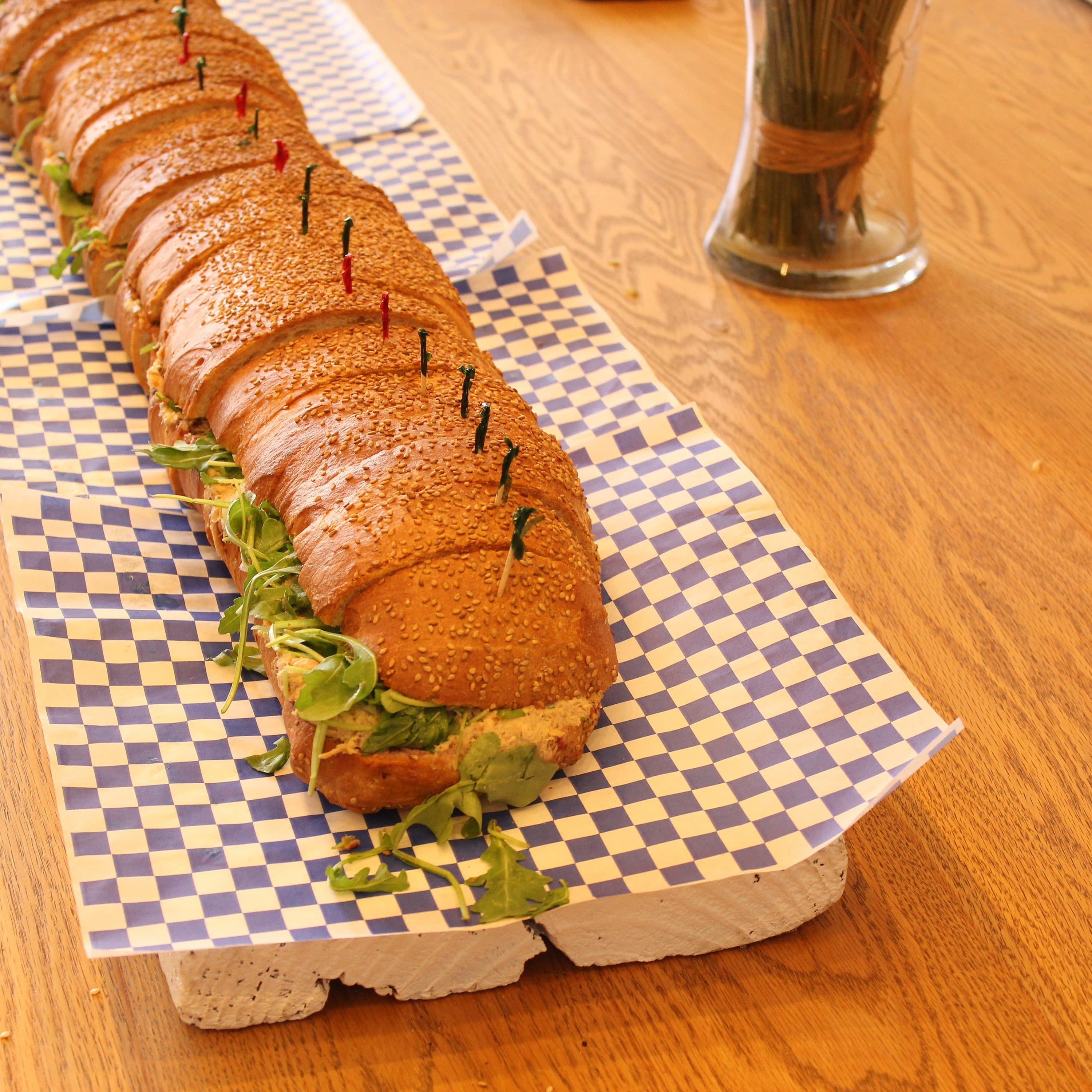 A long sub sandwich topped with sesame seeds is displayed on a wooden table. The sandwich, garnished with fresh greens, is divided into sections using colorful toothpicks. Blue and white checkered paper is placed underneath for decoration.
