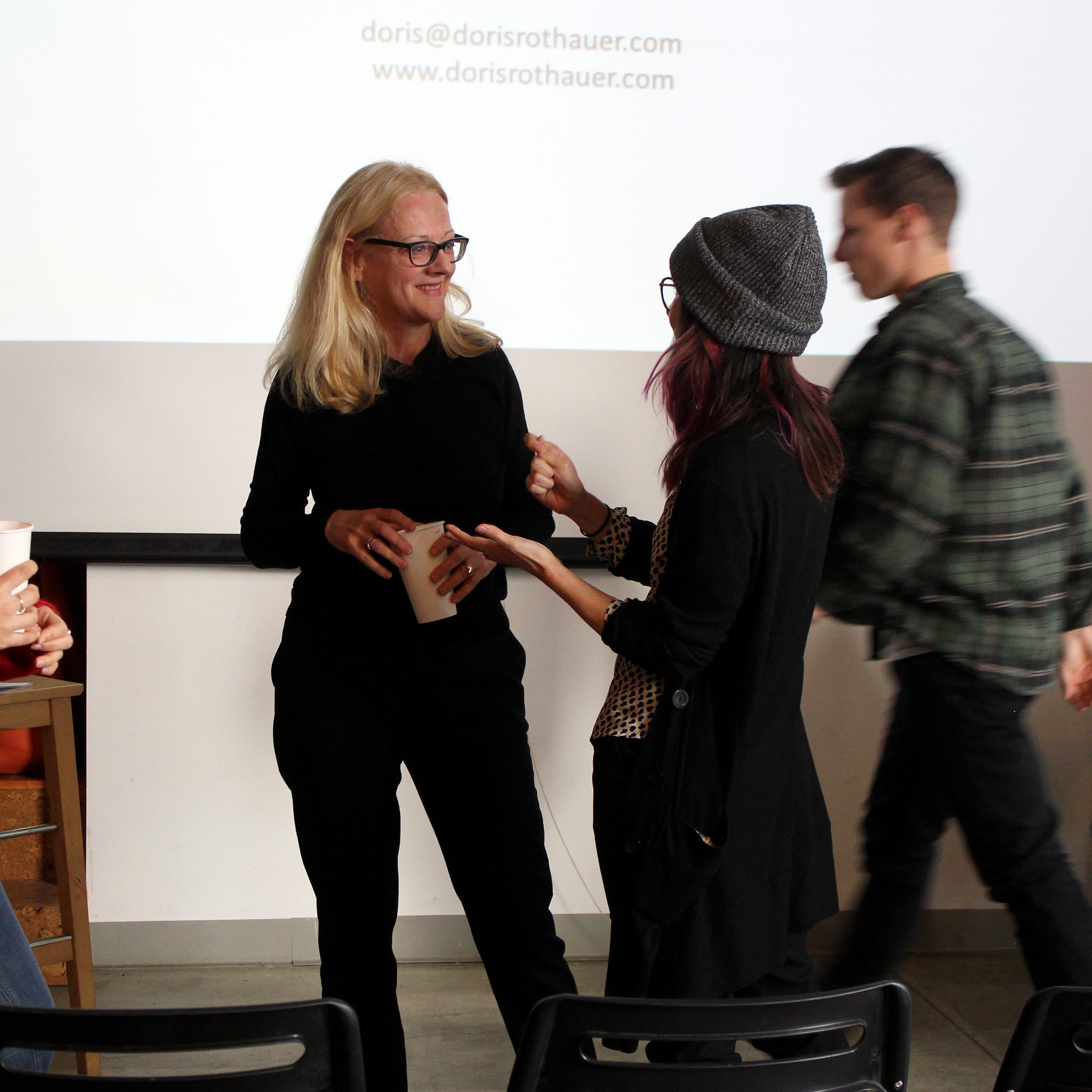 A woman with long blonde hair, wearing glasses and holding a cup, stands talking to another woman with dark hair and a beanie hat. They are indoors with a presentation screen in the background displaying a "Thank You!" message and an email address.