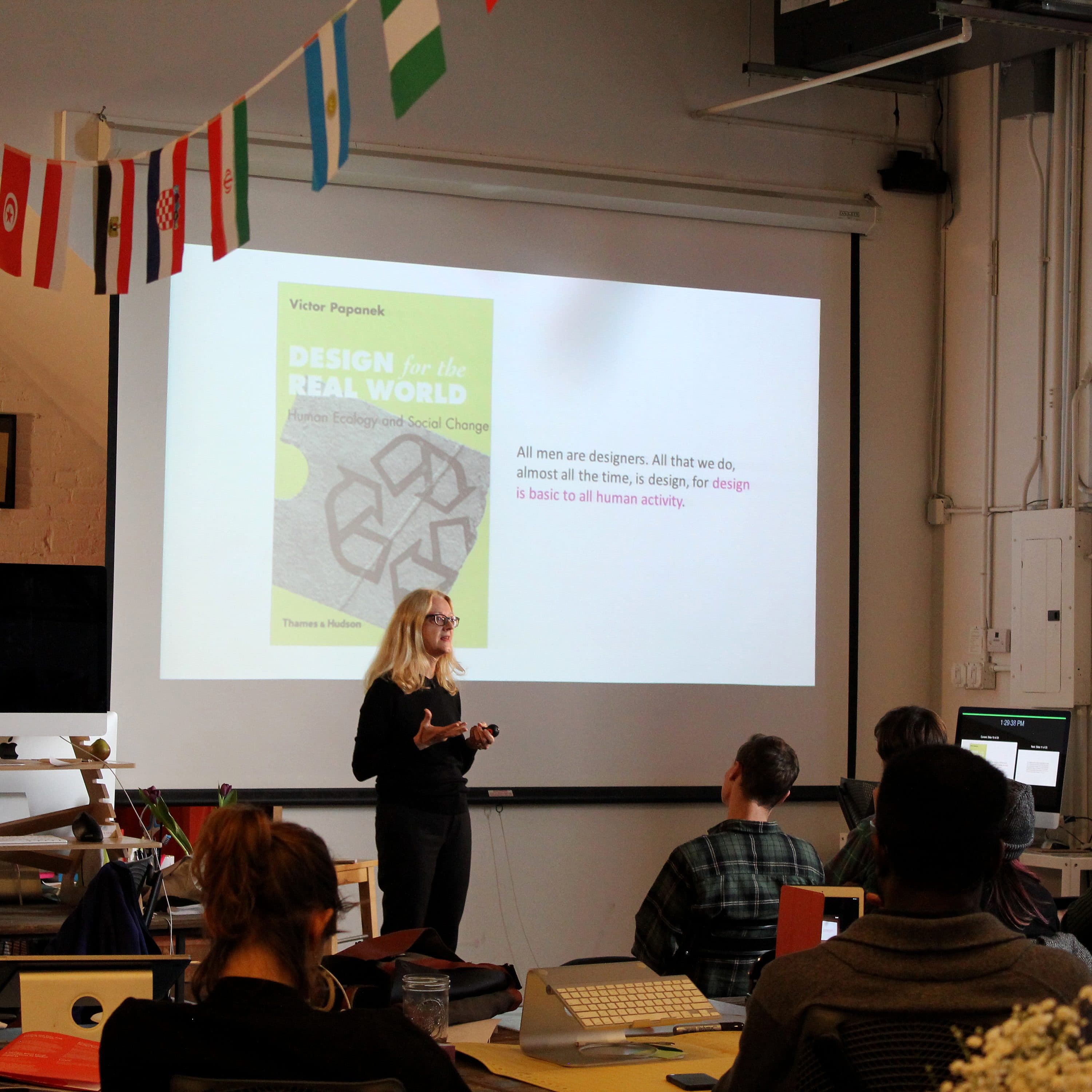 A woman stands in front of a classroom, presenting a slideshow titled "DESIGN for the REAL WORLD" by Victor Papanek. Students are seated, listening attentively. The room is decorated with international flags hanging from the ceiling and various artworks on the walls.