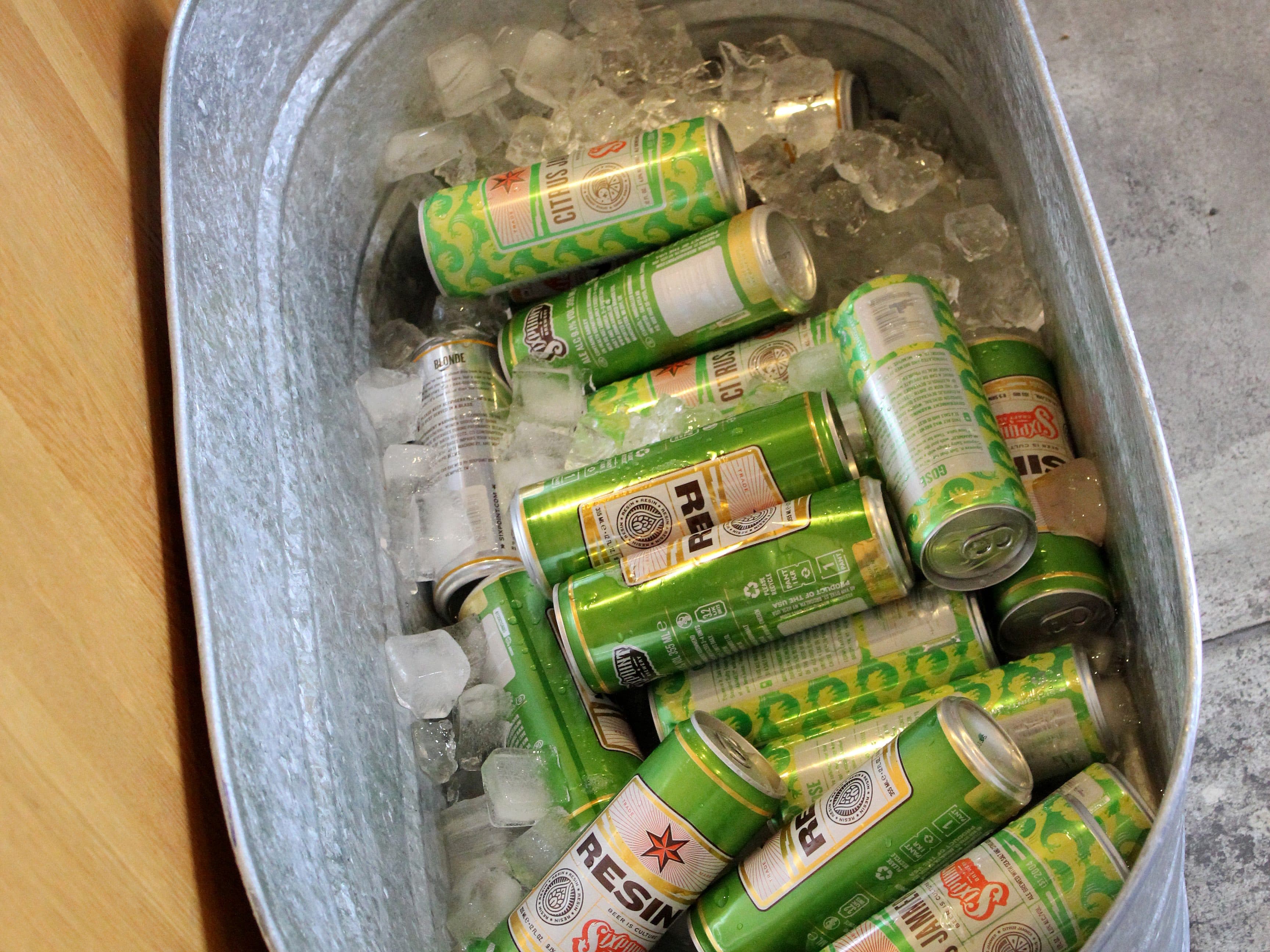 A metal tub filled with ice and various beer cans, primarily featuring green and yellow labels. Several cans are visible, resting in the tub, keeping cool. The setup is on a stone or concrete floor near a wooden surface.