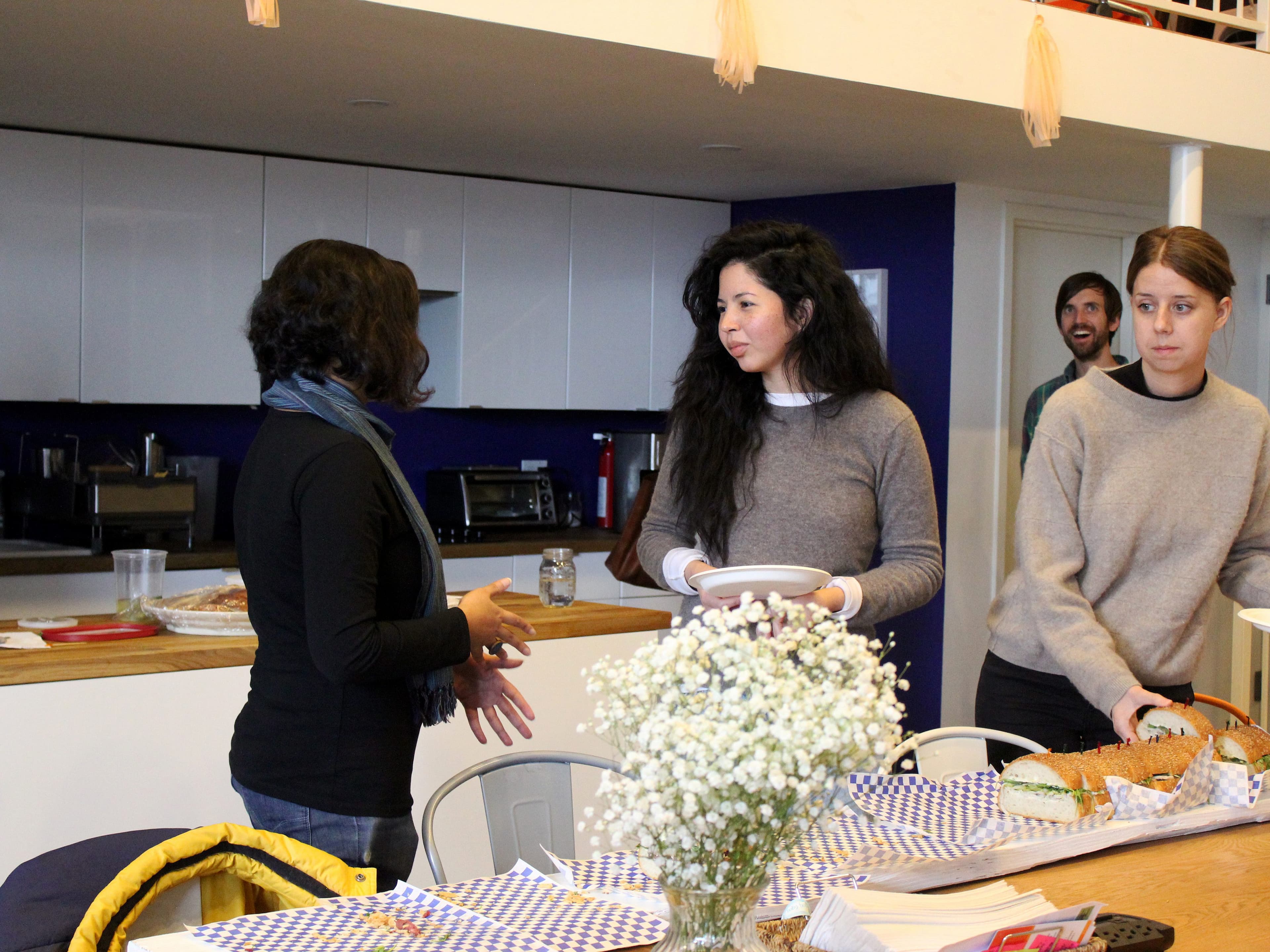Three people are gathered in a kitchen setting with a wooden countertop. Two women are in conversation, holding plates; a man is in the background near a staircase. On the counter are flowers and various food items, including sandwiches and snacks.