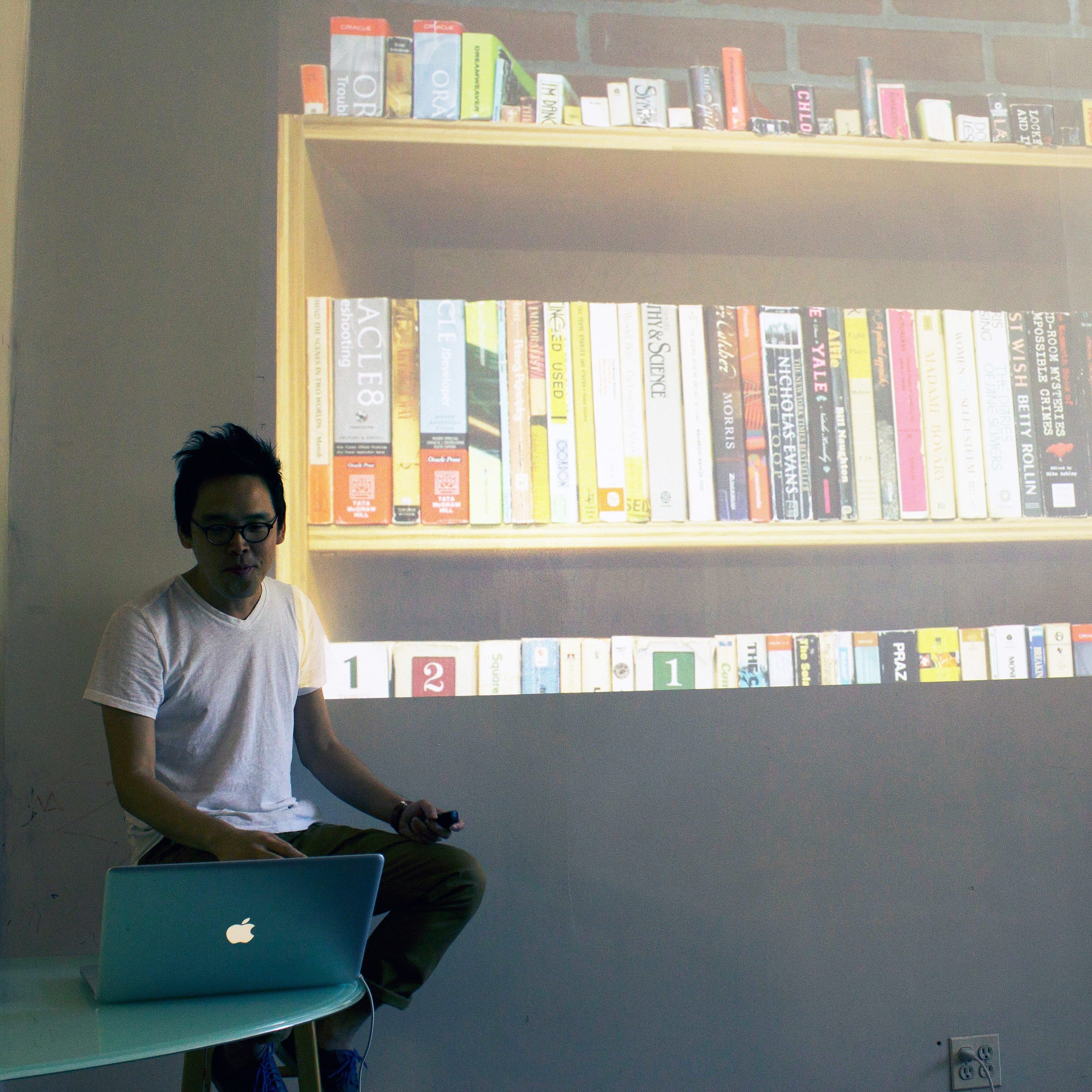 A person wearing glasses and a white T-shirt sits in front of a projected image of a bookshelf full of books. They are holding a device and have a laptop on their lap. The projection is displayed on a large whiteboard behind them.