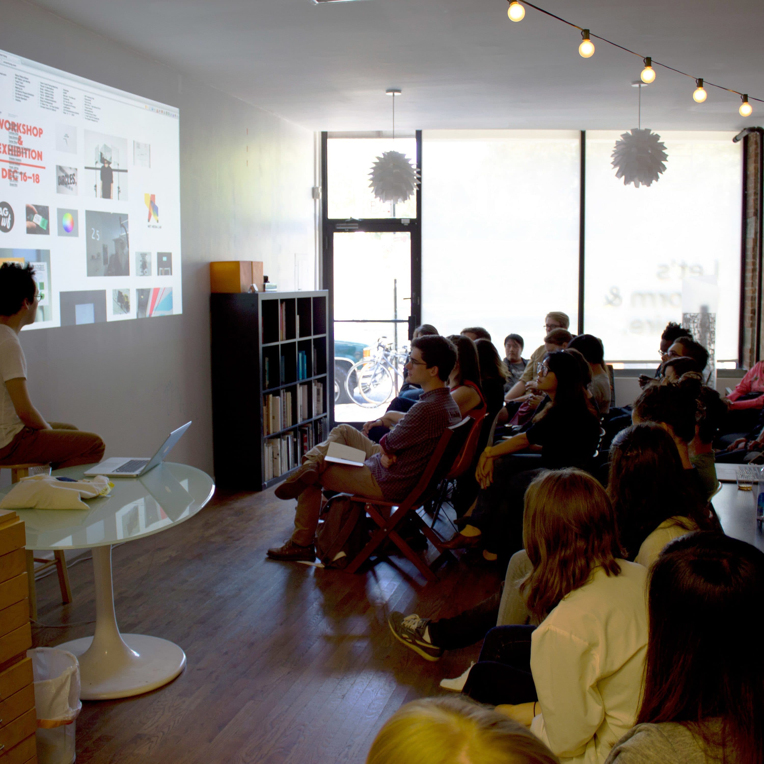 A person is giving a presentation in a room, using a projector to display images and text on a wall. Several people sit in chairs, attentively watching the presentation. The room features a wooden floor, string lights, shelving, and computers on desks.
