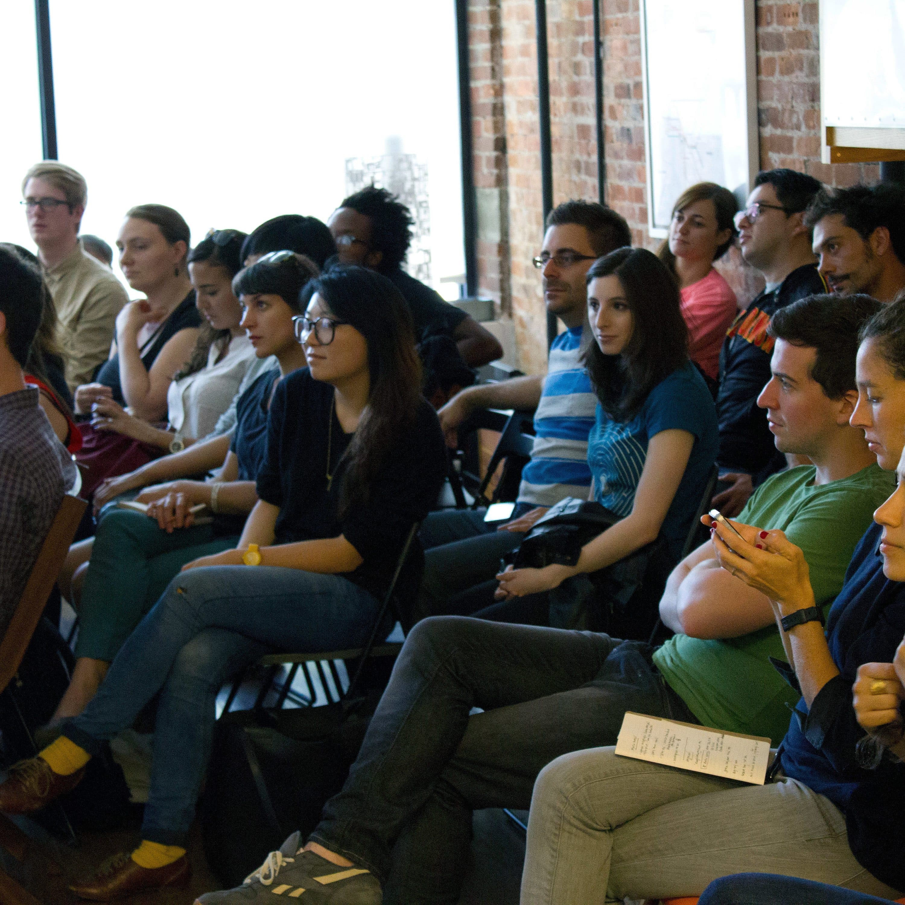 A diverse group of people is seated in a room, attentively listening to a presentation. Some are taking notes or using their phones. The room has large windows and exposed brick walls, creating a casual atmosphere.