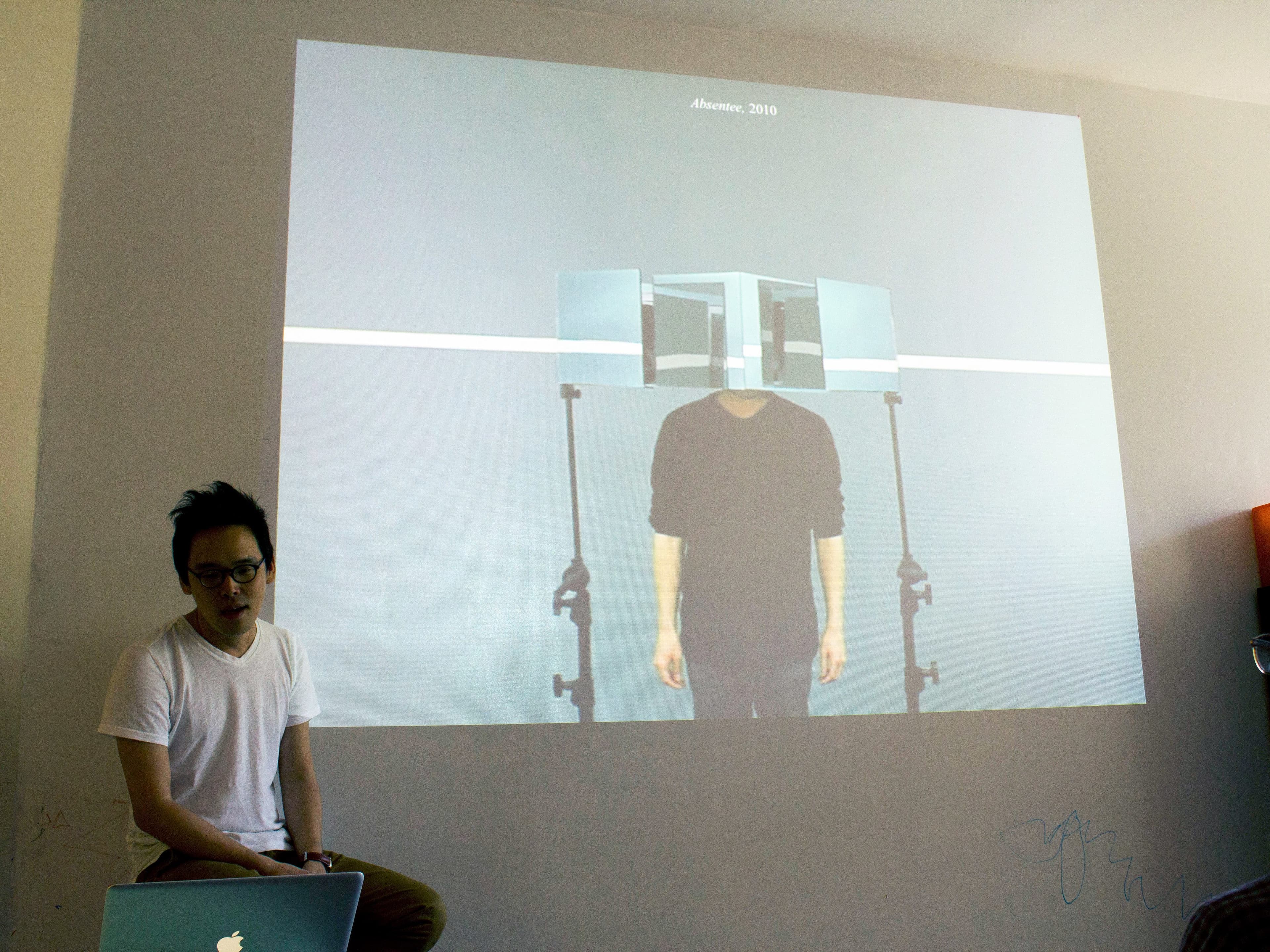 A person in a white t-shirt sits near a laptop, presenting to an audience. Behind them, a large screen displays an art installation featuring a person with square mirrors covering their head. The artwork is titled "Mirrors, 2010.