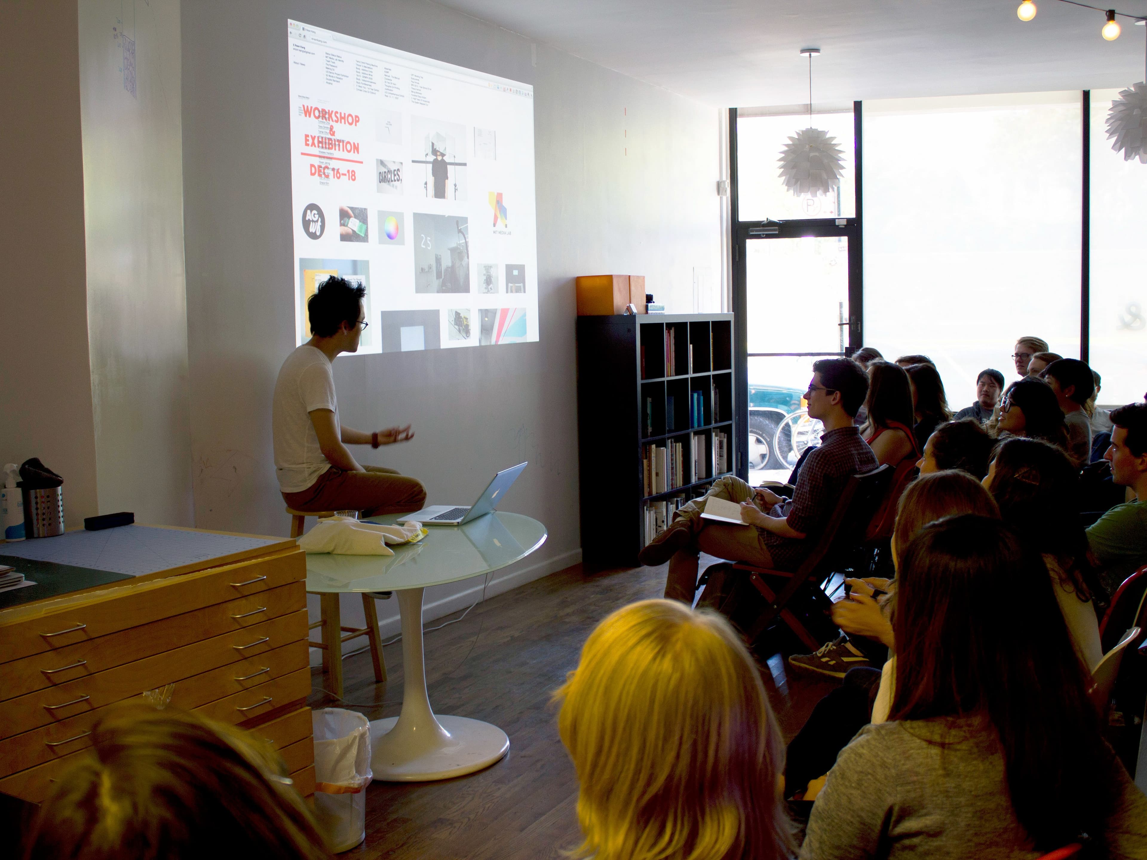A person sits on a table, presenting slides to an attentive audience in a modern, brightly lit room. The slides are projected onto a white wall and include various images and text. The audience consists of people seated in chairs, facing the presenter.