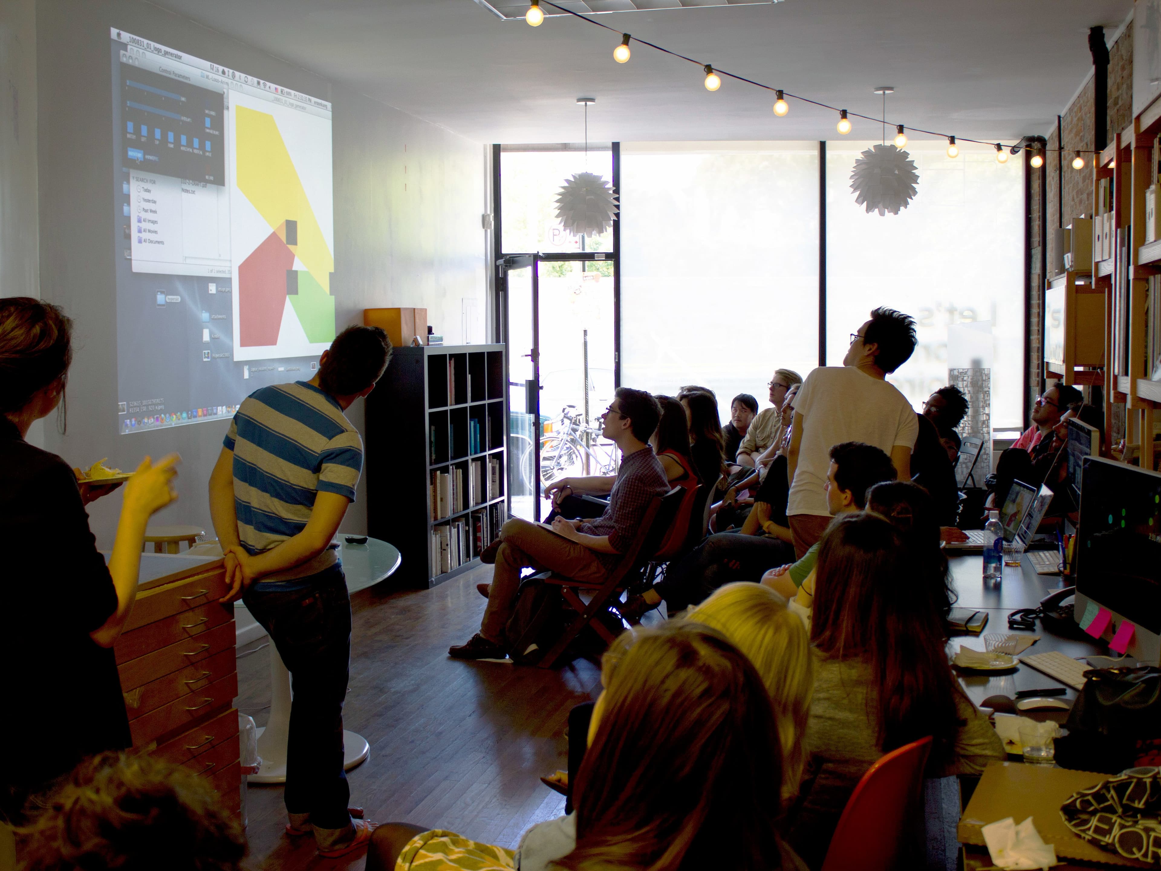 A group of people in a cozy, well-lit room watch a presentation projected on a wall. Some are seated, while others stand near the presenter, who is gesturing at the screen. Shelves with books and a large window are visible in the background.
