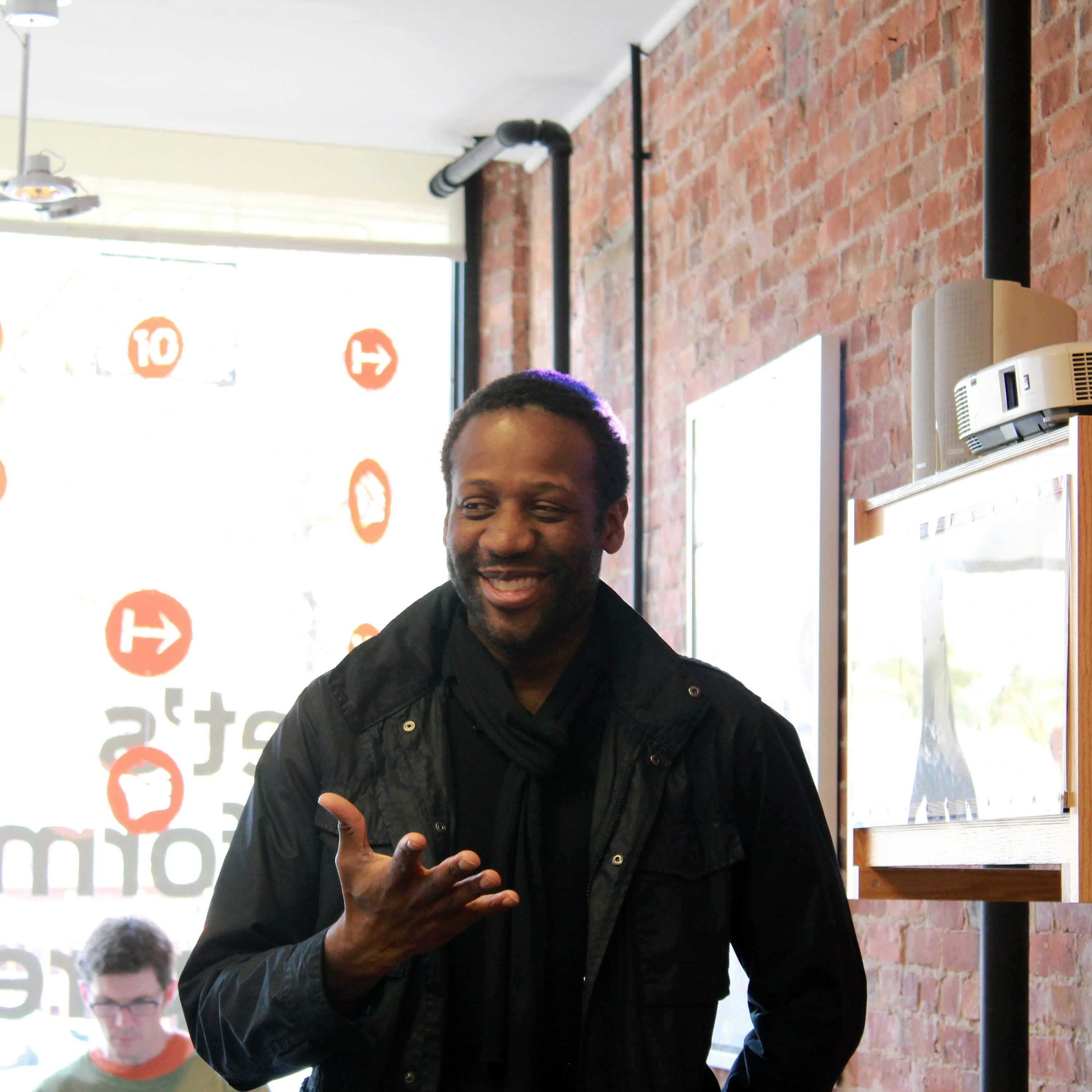 A person in a black jacket and scarf is smiling while giving a speech in a room with exposed brick walls. There are shelves, a window displaying a logo with orange symbols, and a person with glasses in the background.