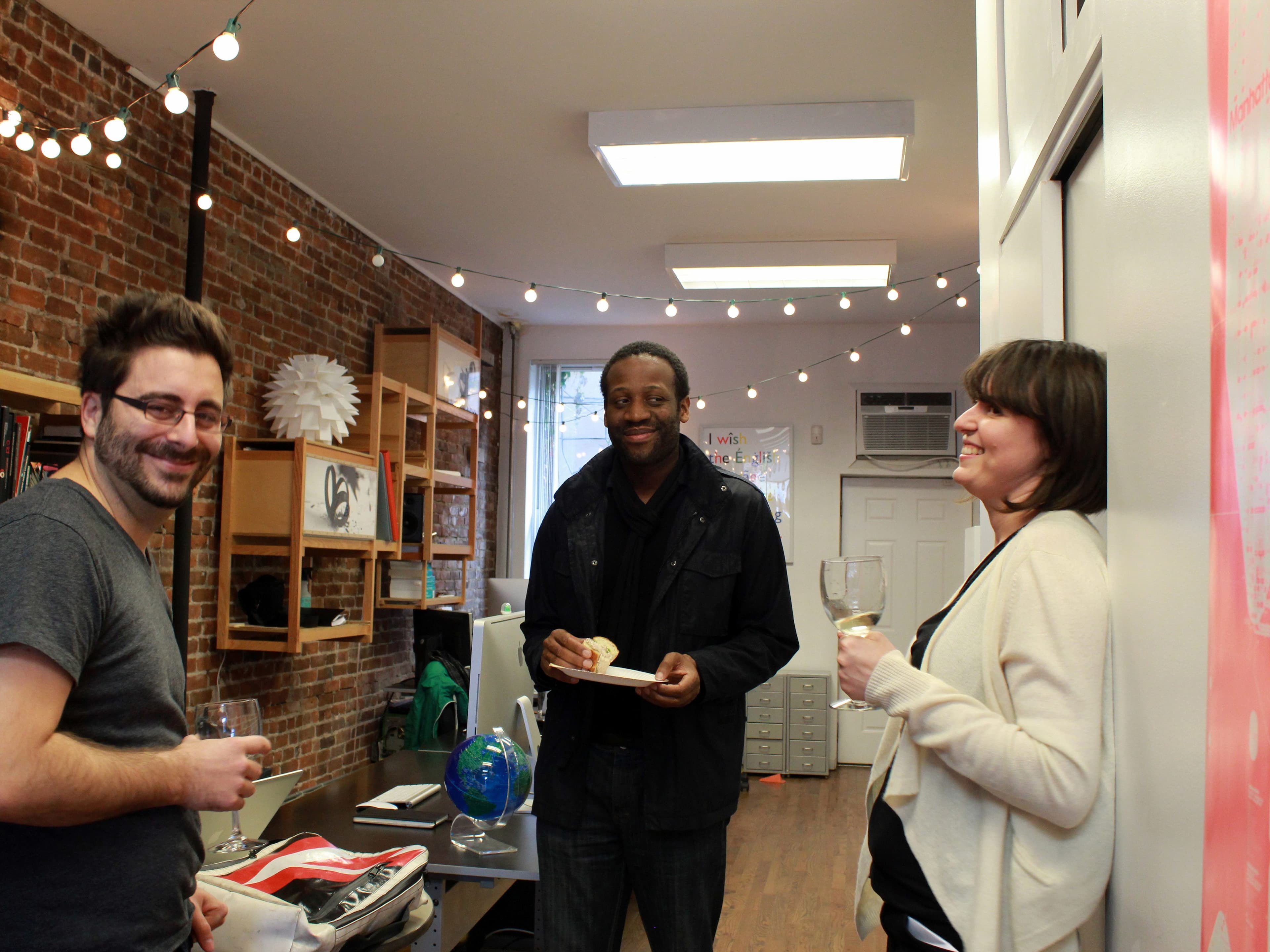 Three people are socializing in a cozy office with string lights and exposed brick walls. Two men and one woman are smiling and holding drinks, with one man holding a plate. Shelving in the background displays various items, including a globe.