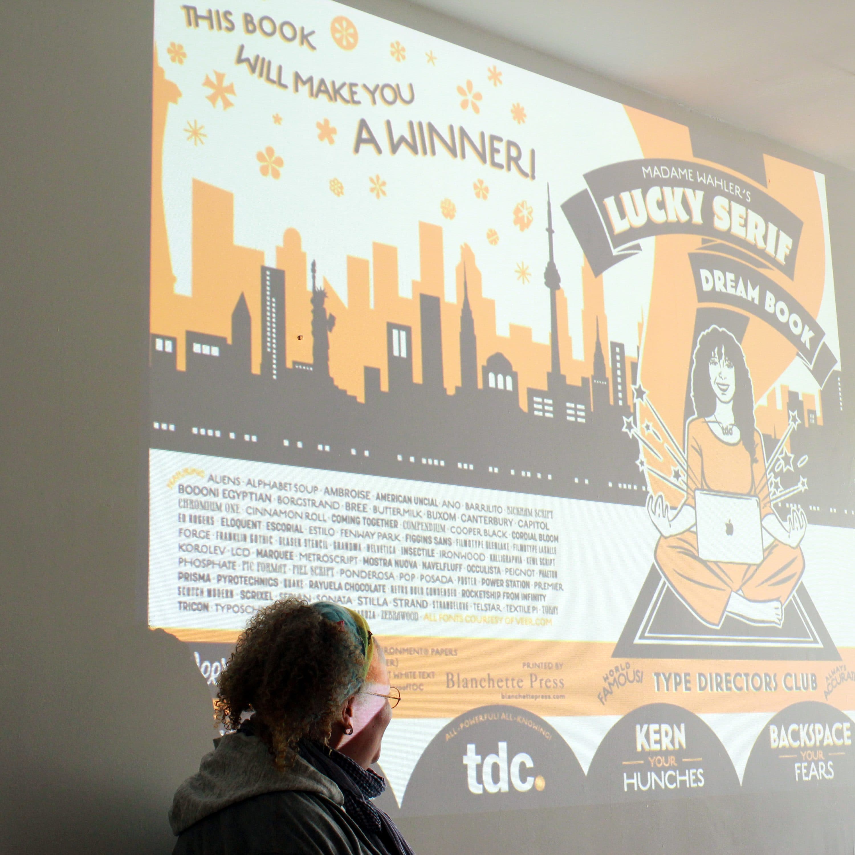 A person with curly hair is pointing at a projected image on a white wall. The image is a promotional poster in orange, black, and white for a book titled "Madame Hample's Lucky Serif Dream Book," featuring a stylized cityscape and a seated figure with a laptop.