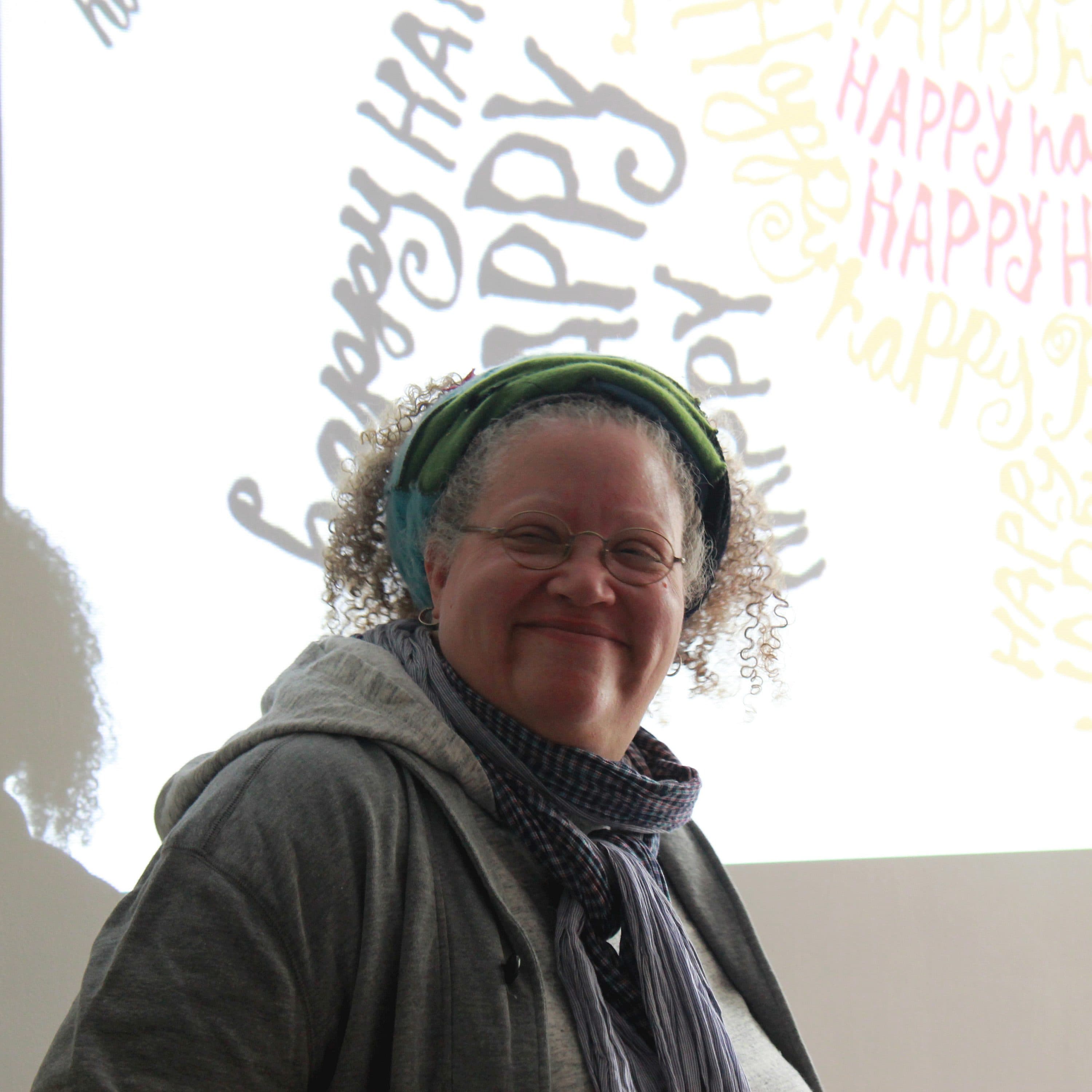 A person with curly hair, wearing glasses and a colorful headscarf, smiles while standing in front of a wall projection displaying the word "HAPPY" multiple times in various fonts and colors. The person is dressed in light-colored attire and a striped scarf.