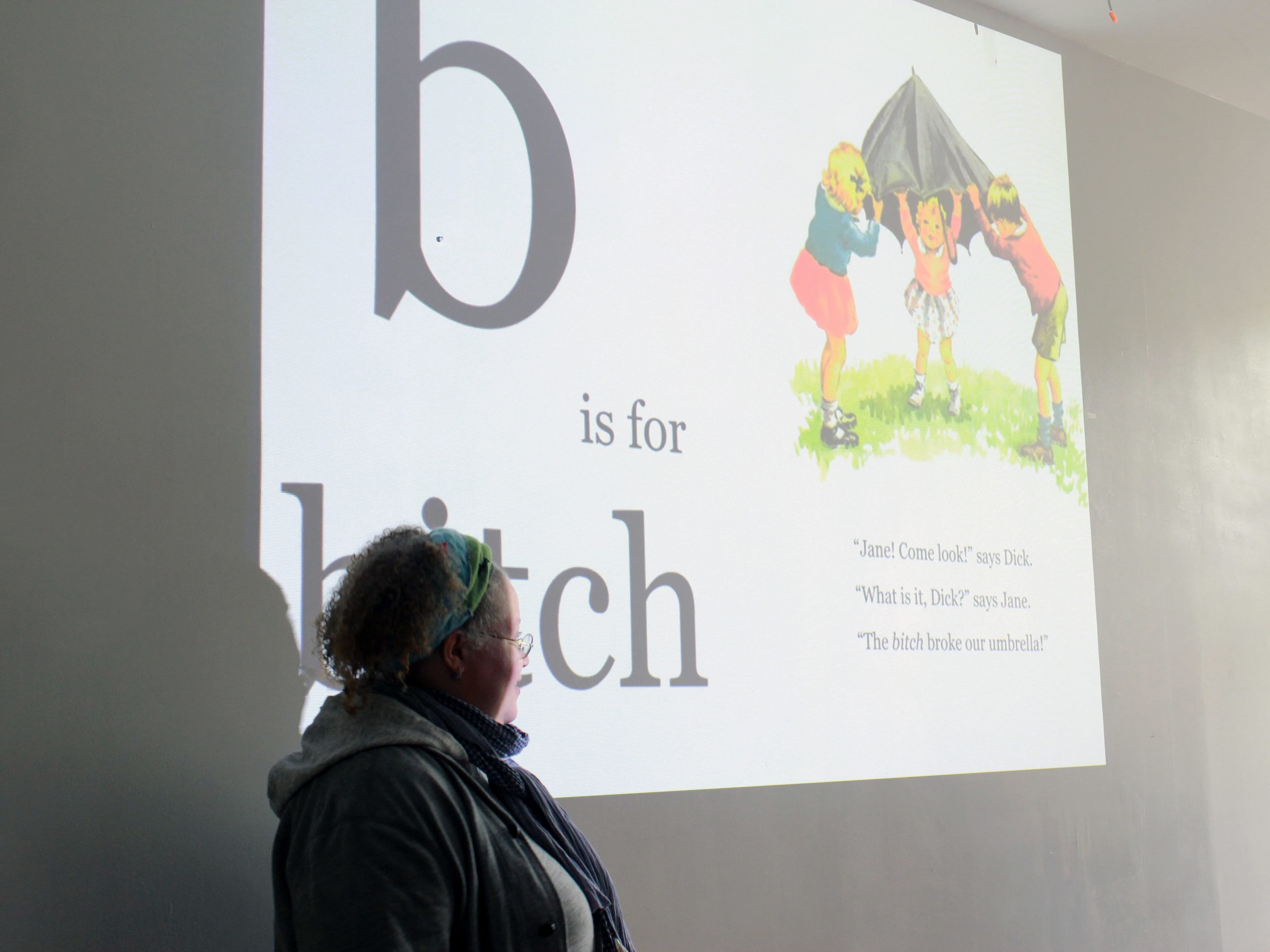 A person stands in front of a projection on a wall displaying a page from a book. The page shows an illustration of children playing underneath a blanket with the text "b is for bitch" and a conversation between characters about their umbrella.