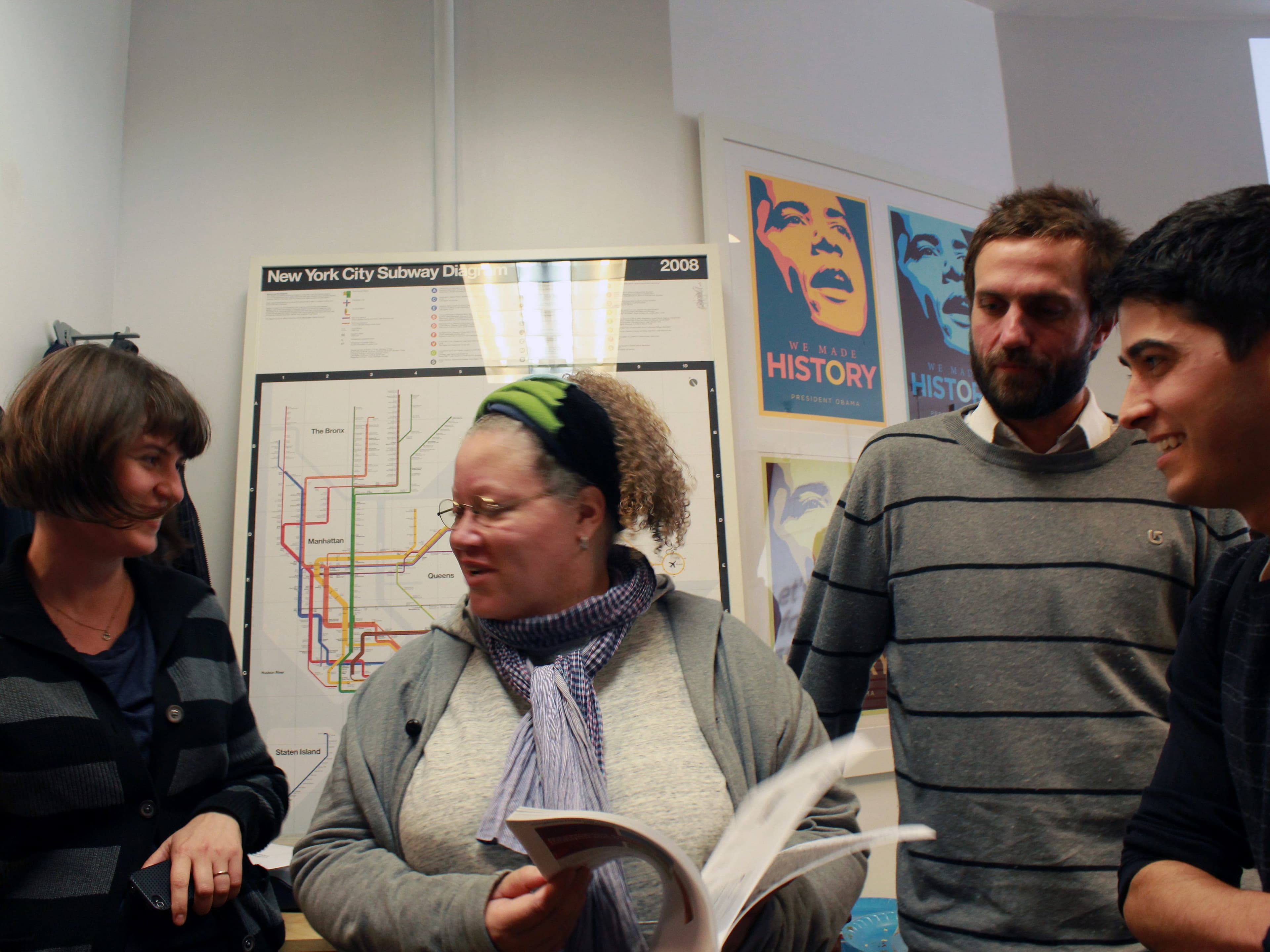 Four people are standing in a room engaged in conversation. One person is holding a book or magazine. A New York City Subway map and posters with bold designs are visible on the wall behind them. They appear to be in an office or informal setting.