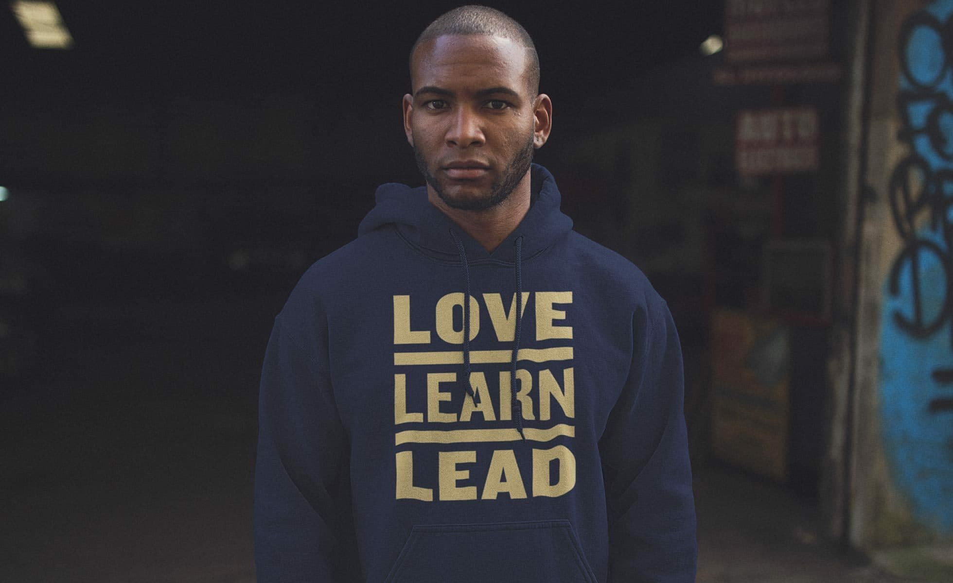 A person stands facing the camera wearing a navy blue hoodie with the words "LOVE LEARN LEAD" printed in bold yellow text. The background is dark and features graffiti on the right side.