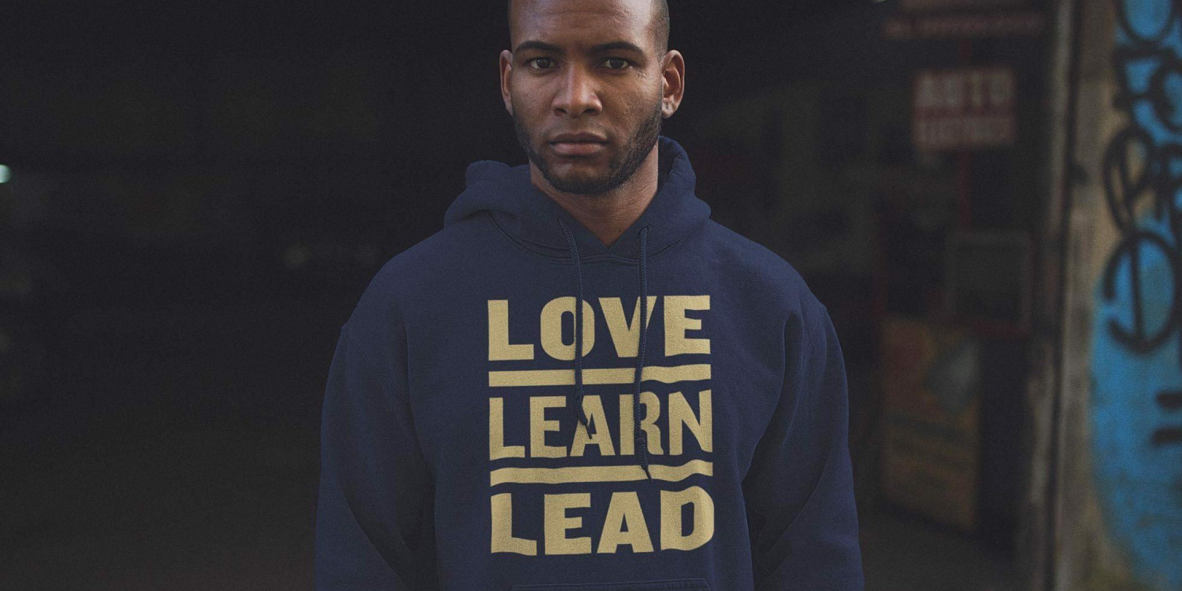 A person stands facing the camera wearing a navy blue hoodie with the words "LOVE LEARN LEAD" printed in bold yellow text. The background is dark and features graffiti on the right side.