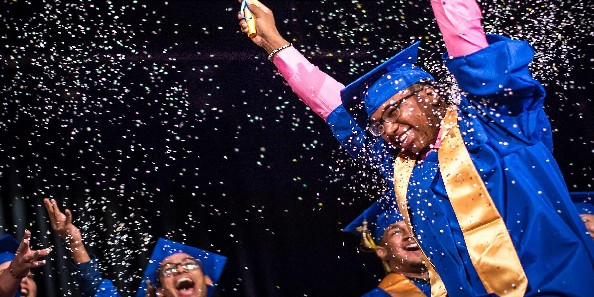 A group of graduates in blue caps and gowns celebrate on stage. One person in the front raises both arms joyfully, with one hand holding a confetti popper, releasing a shower of confetti. The background is filled with other graduates smiling and cheering.