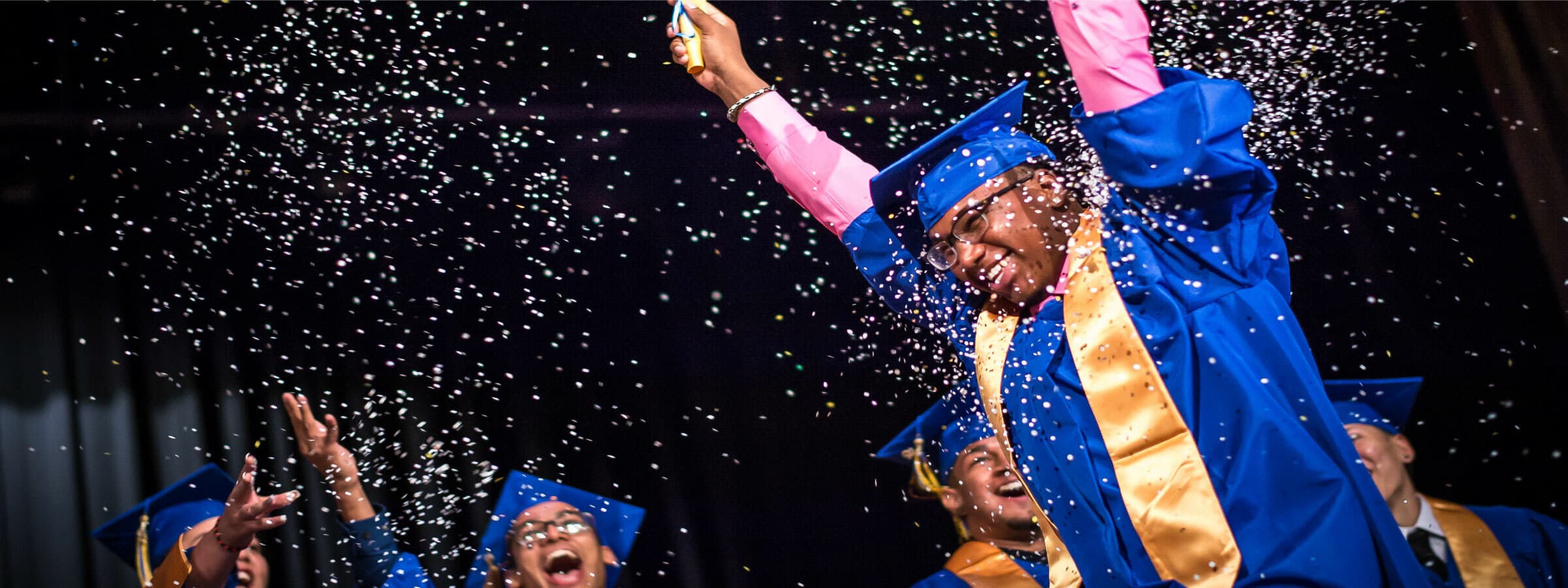 A group of graduates in blue caps and gowns celebrate on stage. One person in the front raises both arms joyfully, with one hand holding a confetti popper, releasing a shower of confetti. The background is filled with other graduates smiling and cheering.