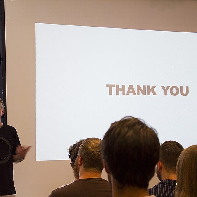 A man stands in front of a group of people, giving a presentation. He is next to a large screen that displays the words "THANK YOU." The audience is sitting and facing the presenter.