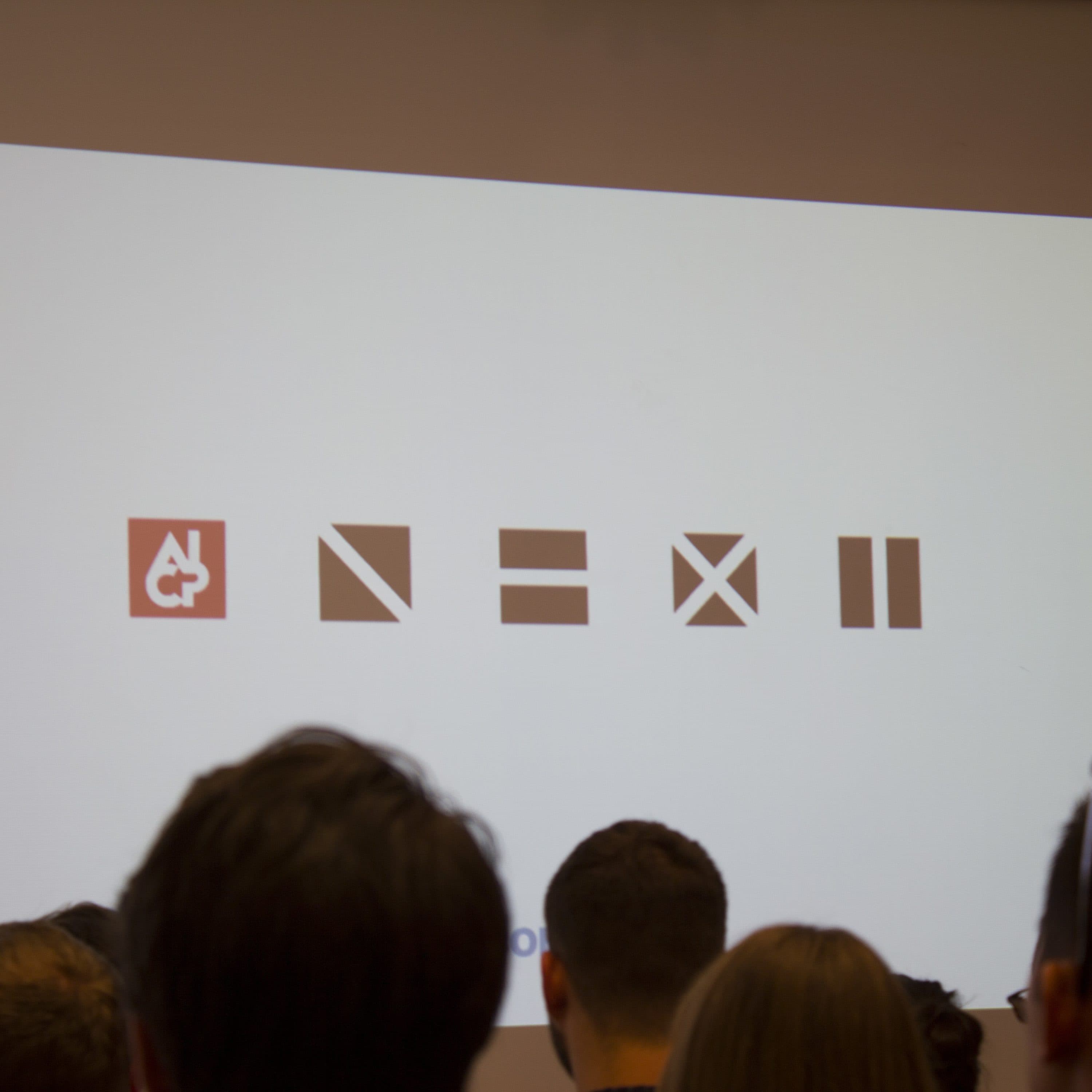 An audience watches a presentation showing various symbols on a screen. From left to right, the symbols are: a red square with a white abstract figure, a black diagonal line, an equal sign, a black X, and two black vertical lines denoting pause.