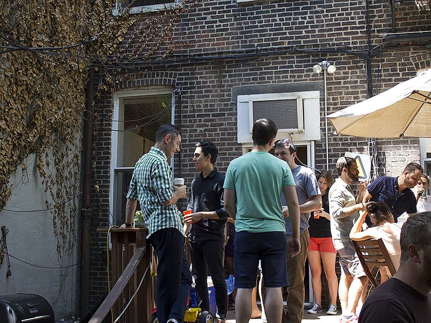 A group of people are gathered outdoors at a backyard social event. Some are standing and chatting while holding drinks, and others are sitting around a table. The scene is set against a brick building and partially covered with an umbrella.