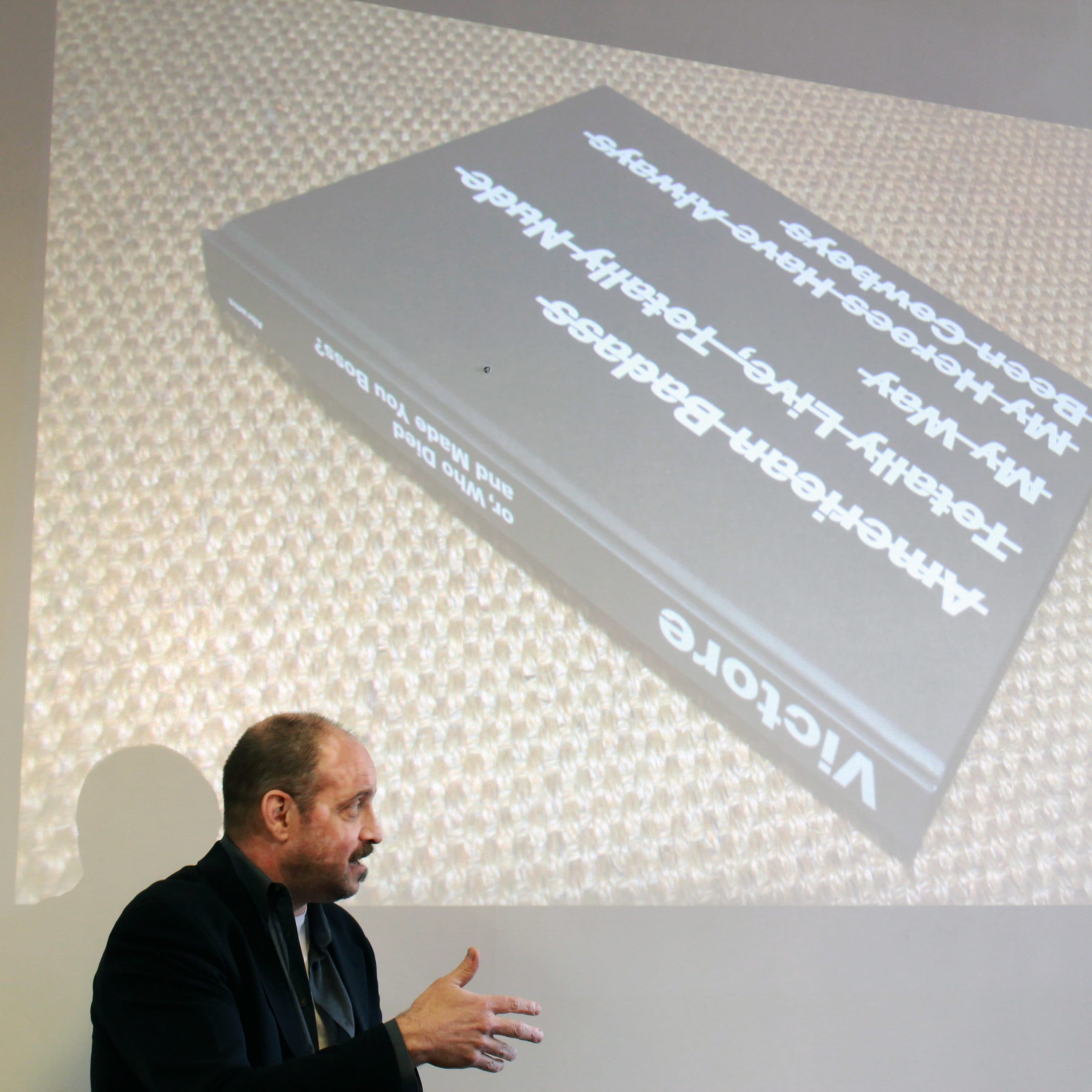 A man in a suit gestures while speaking in front of a projected image of a book titled "American B ALBB Moscow/ ViktoRE Family Tree Branch City History" on a wall. The book is black with white text and is placed on a textured surface.