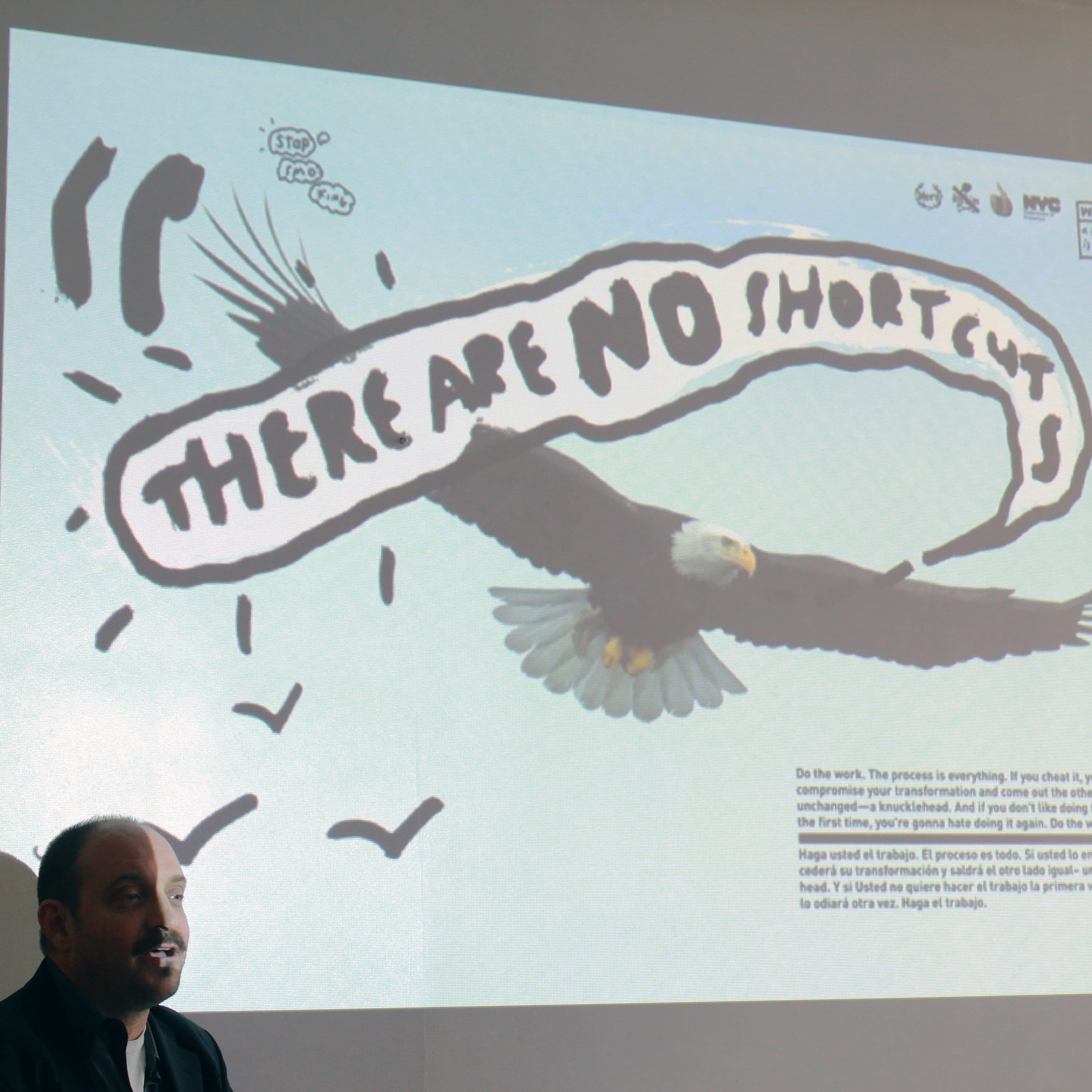 A man in a suit stands in front of a projection of an illustrated bald eagle flying with the text, "THERE ARE NO SHORTCUTS" in a speech bubble. Below, smaller text is visible but illegible from this distance. The background is a plain white wall.