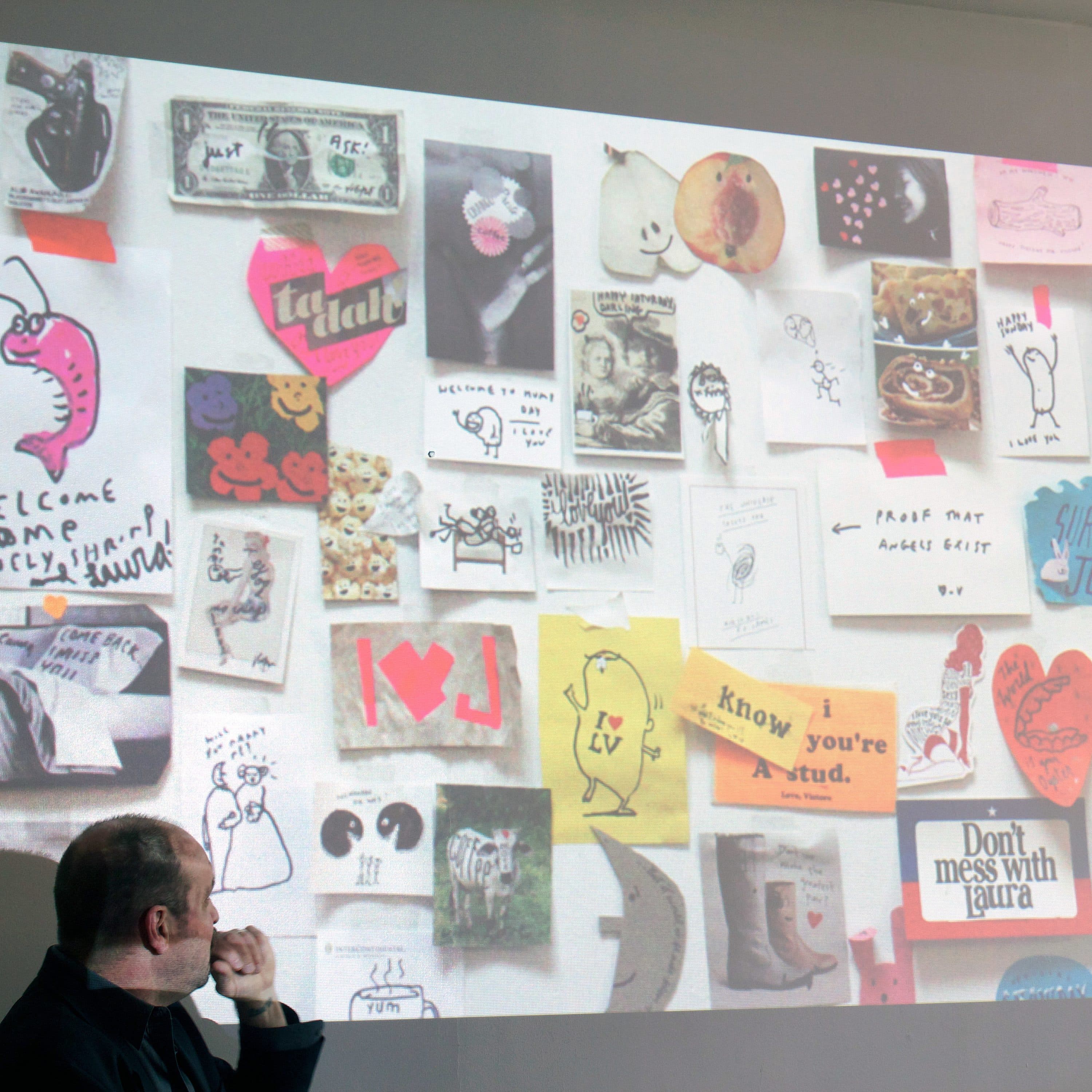 A person is sitting and looking at a wall filled with various drawings, notes, and pictures. The collage includes doodles, heart shapes, messages, and other artistic elements. The person appears to be contemplating or analyzing the colorful display.