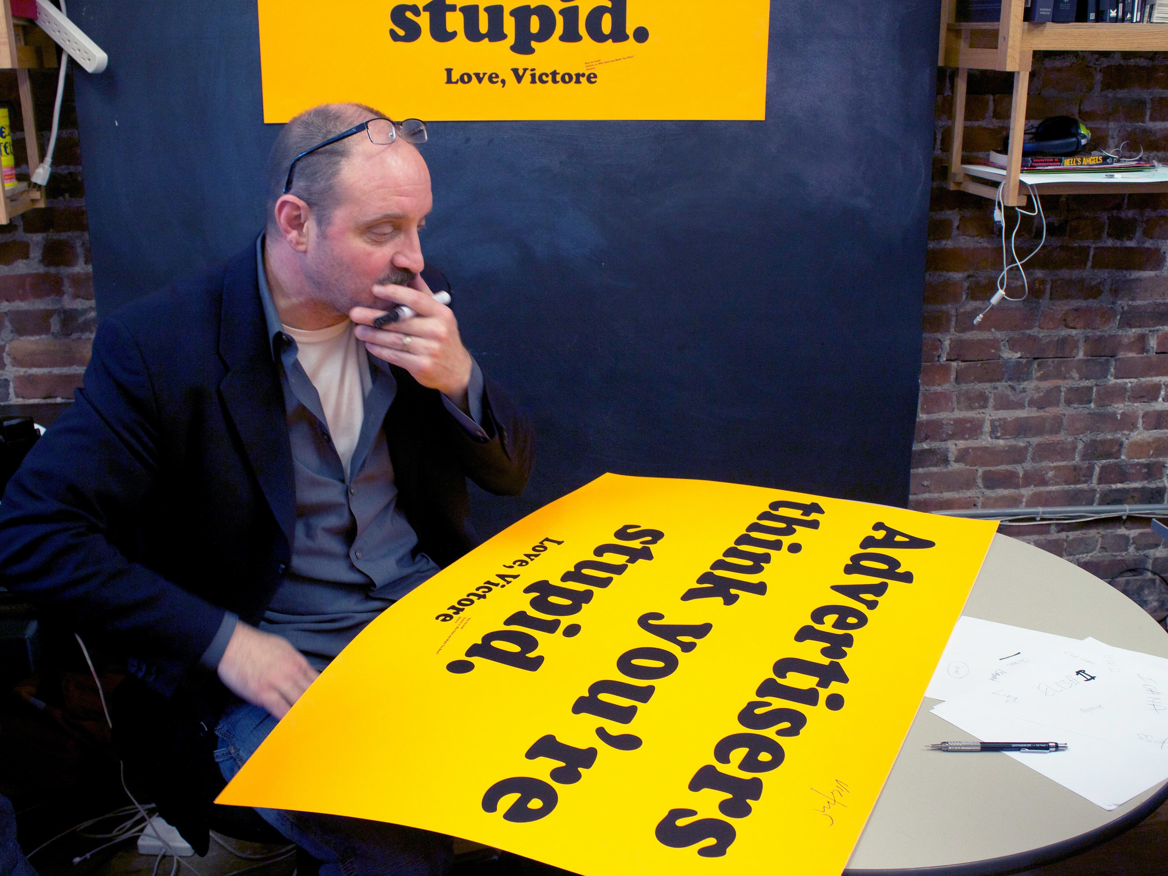 A man sits at a table and holds a marker to his mouth while looking at a large yellow poster with black text that reads, "Advertisers think you're stupid. Love, Victore." A second yellow poster with the same message is displayed on a blackboard behind him.
