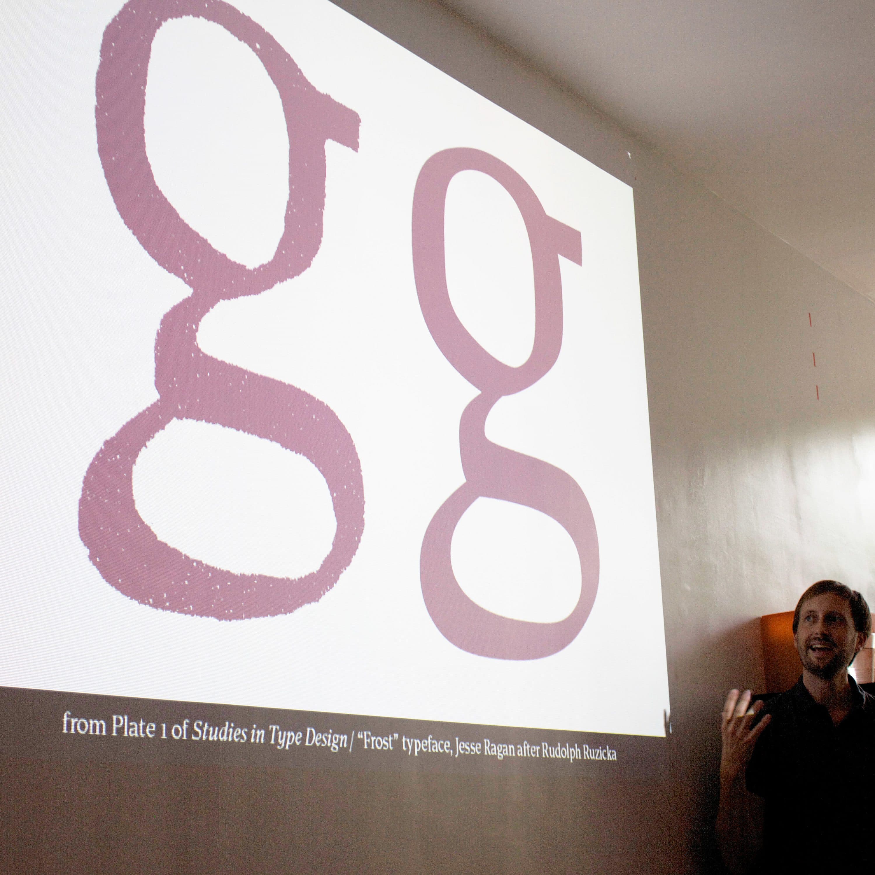 A person stands in front of a projection showing two typeface styles of the letter "g" on a white wall. The slide text reads, "from Plate 1 of Studies in Type Design,” with additional details about the typeface. The person is speaking and gesturing with one hand.