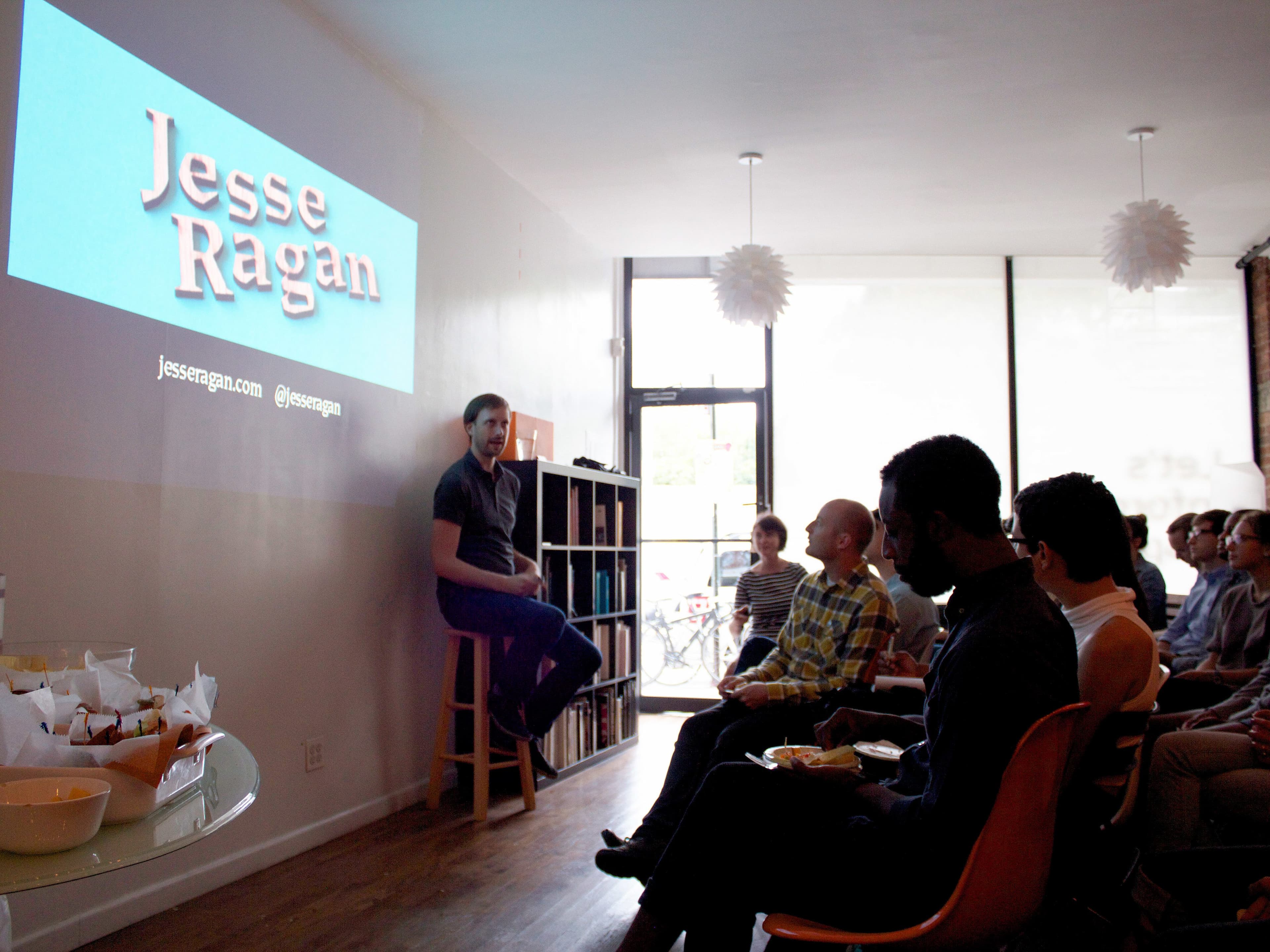 A man sits on a stool in front of a small audience in a brightly lit room, giving a presentation. The screen next to him displays "Jesse Ragan" along with a web address and a Twitter handle. Snacks and drinks are visible on a table to the left.