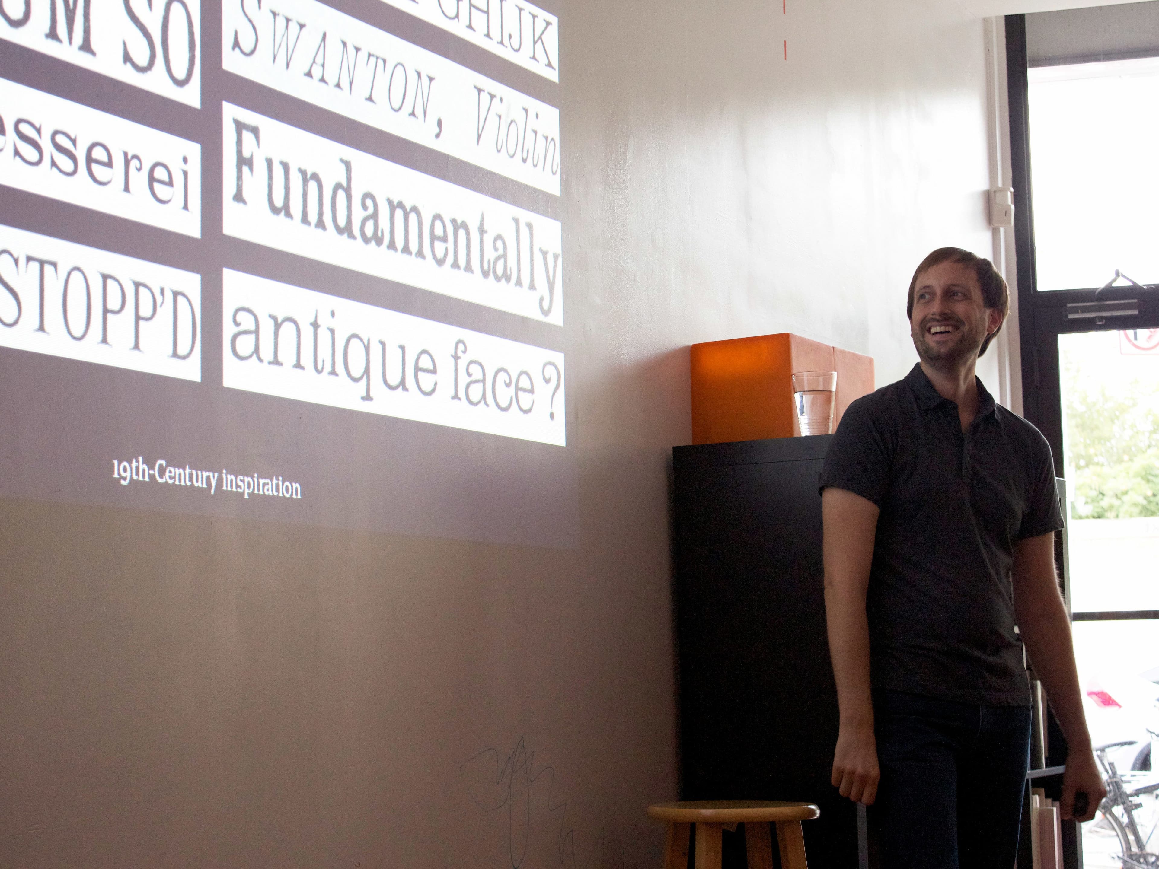 A man in a black shirt stands smiling, looking at a projection on the wall that displays text in various fonts and styles, including phrases like "Fundamentally antique face?" and "19th-Century Inspiration." He stands next to a stool and a dark cabinet with a glass of water.