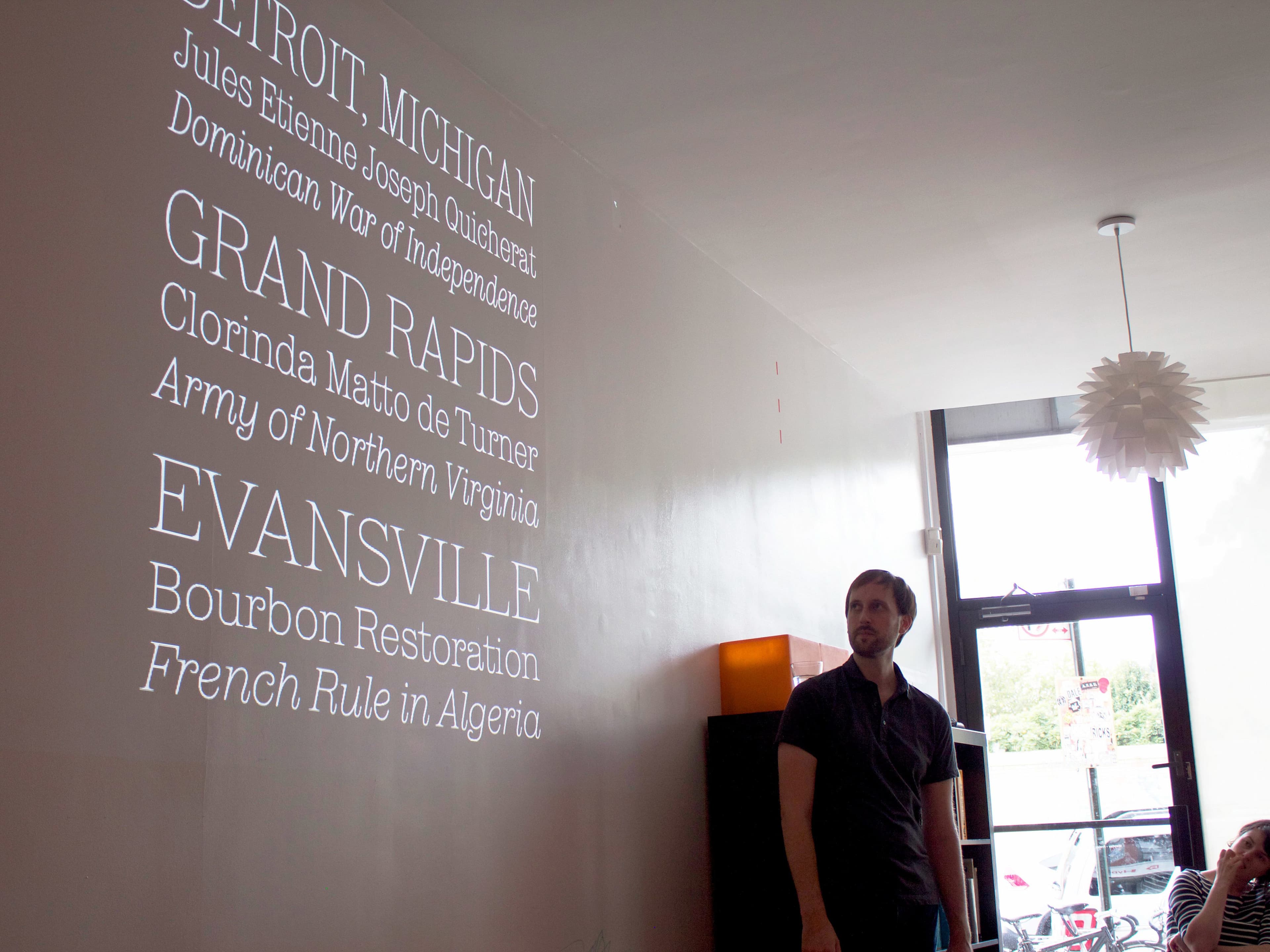 A man, standing in a room with a modern ceiling light fixture, looks up at projected text on the wall. The text includes locations such as Detroit, Michigan, and Grand Rapids, along with historical references like the Dominican War of Independence and French Rule in Algeria.