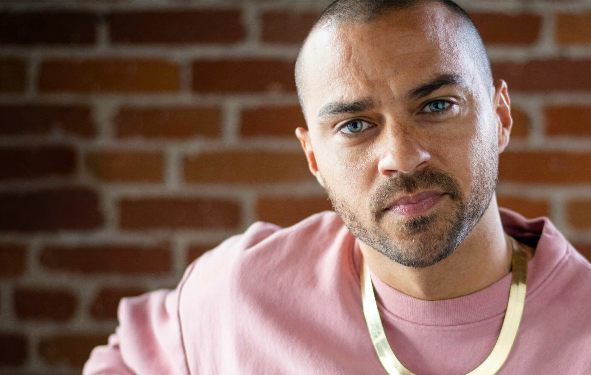 A person with short hair, a beard, and blue eyes is wearing a pink shirt and a gold necklace. They are looking directly at the camera with a brick wall as the background.