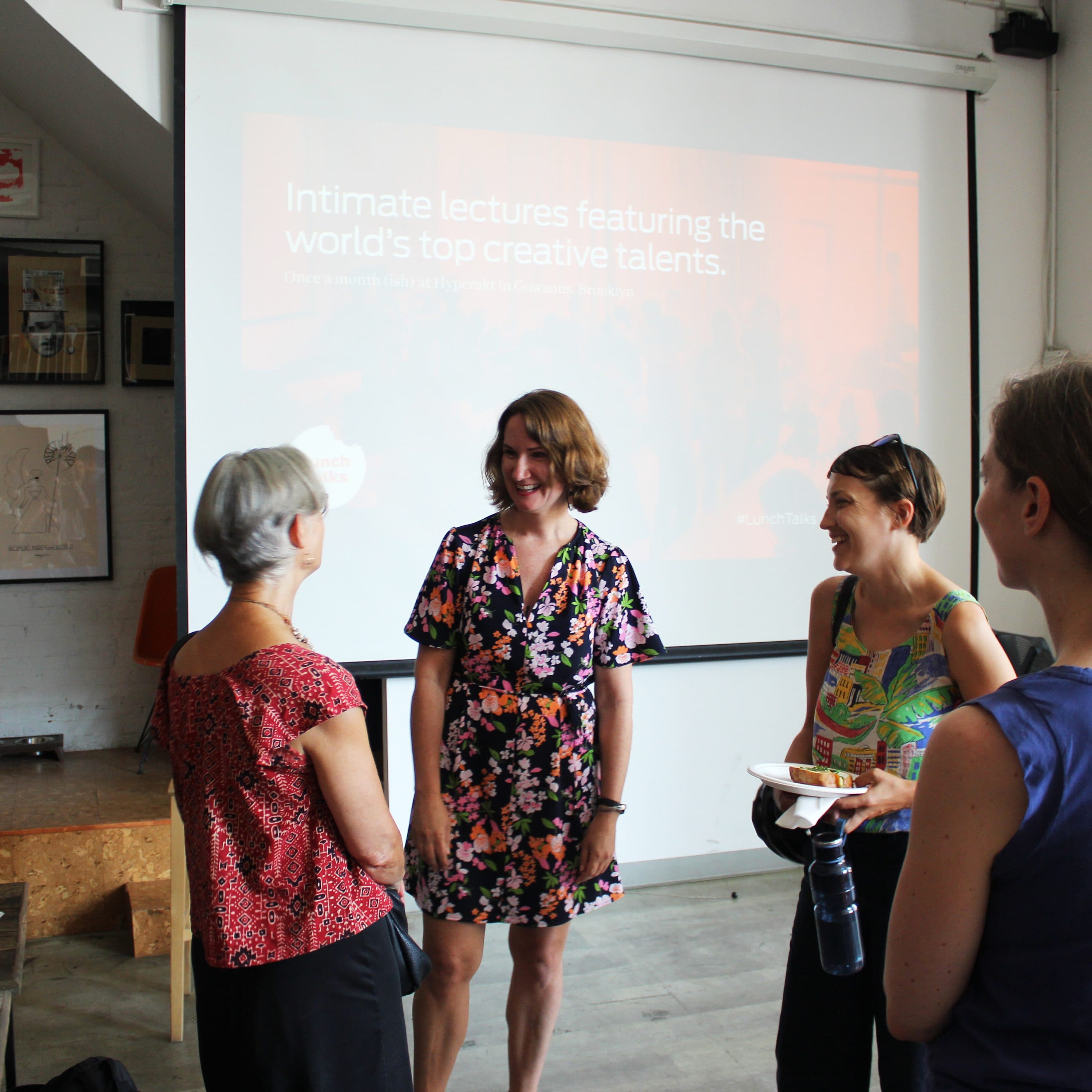 Four women are chatting and smiling in a casual indoor setting with art on the wall and a projection screen in the background. The screen displays the text: "Intimate lectures featuring the world's top creative talents." One woman is holding a camera.