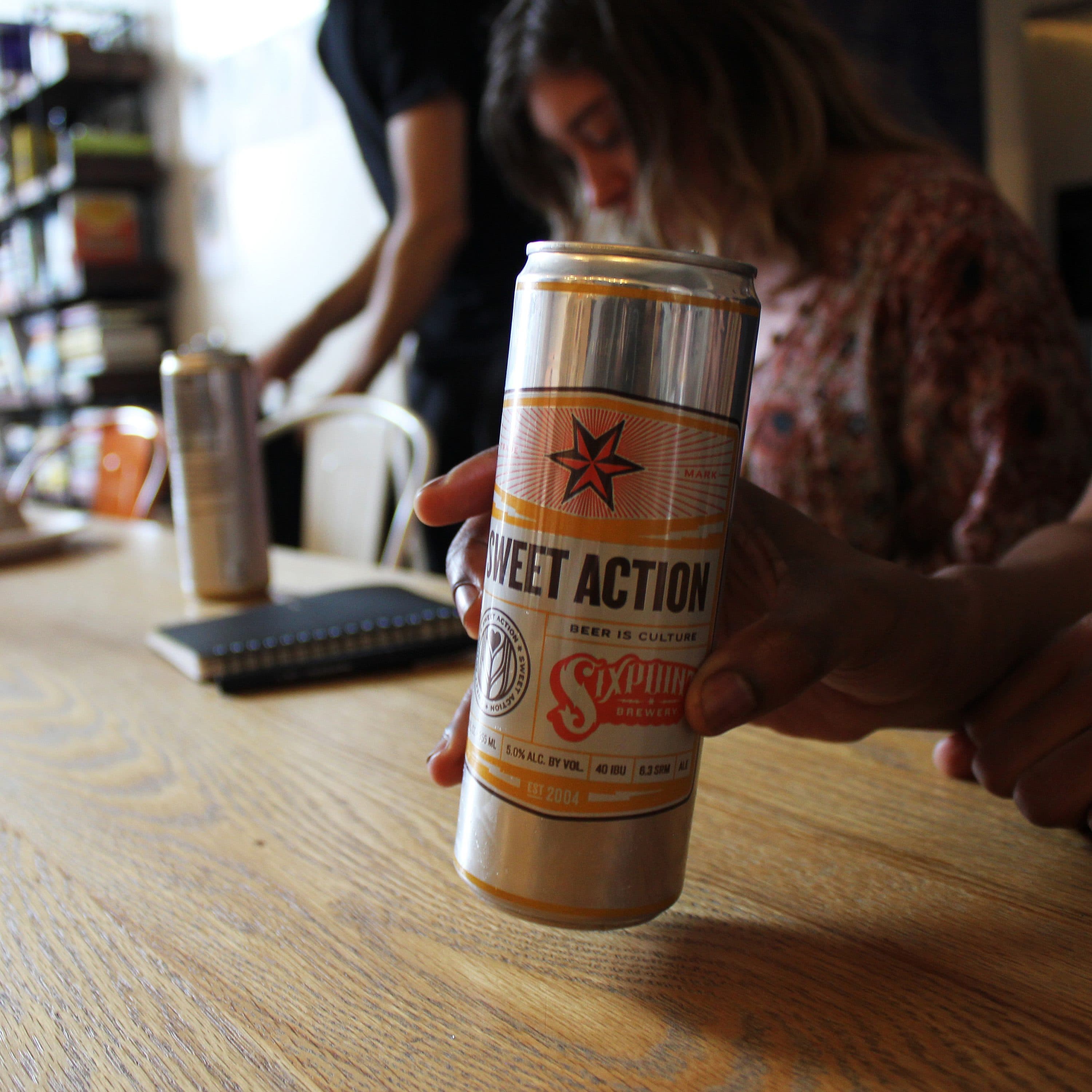 A person holds a can of Sixpoint Brewery's Sweet Action beer over a wooden table. In the background, two other people are partially visible—one seated and the other standing. The scene is indoors, with bookshelves and a refrigerator in the background.