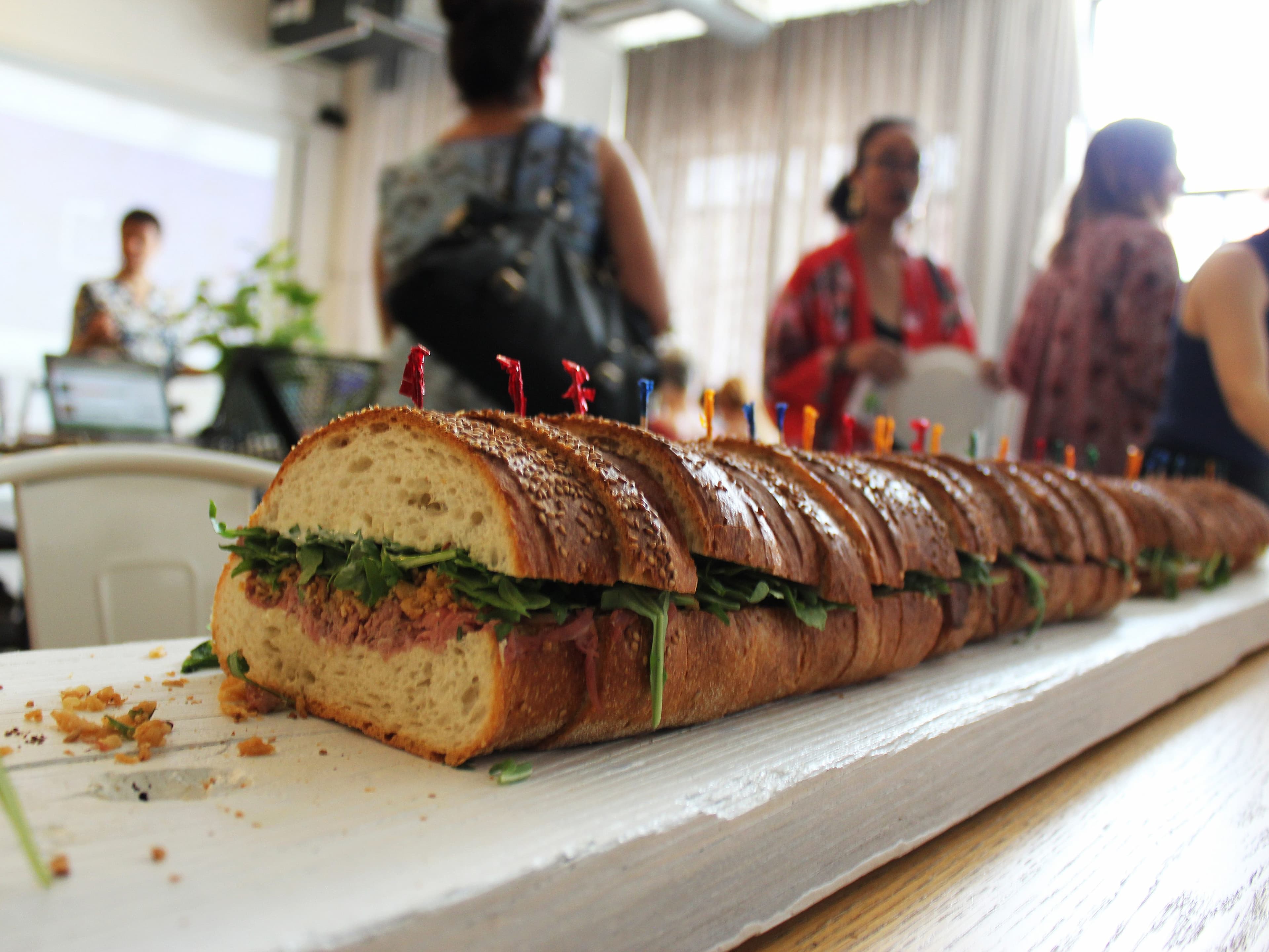 A long sandwich cut into many pieces, with visible greens and packed filling, placed on a white wooden board. In the background, there are several people engaging in conversation in a bright indoor setting.