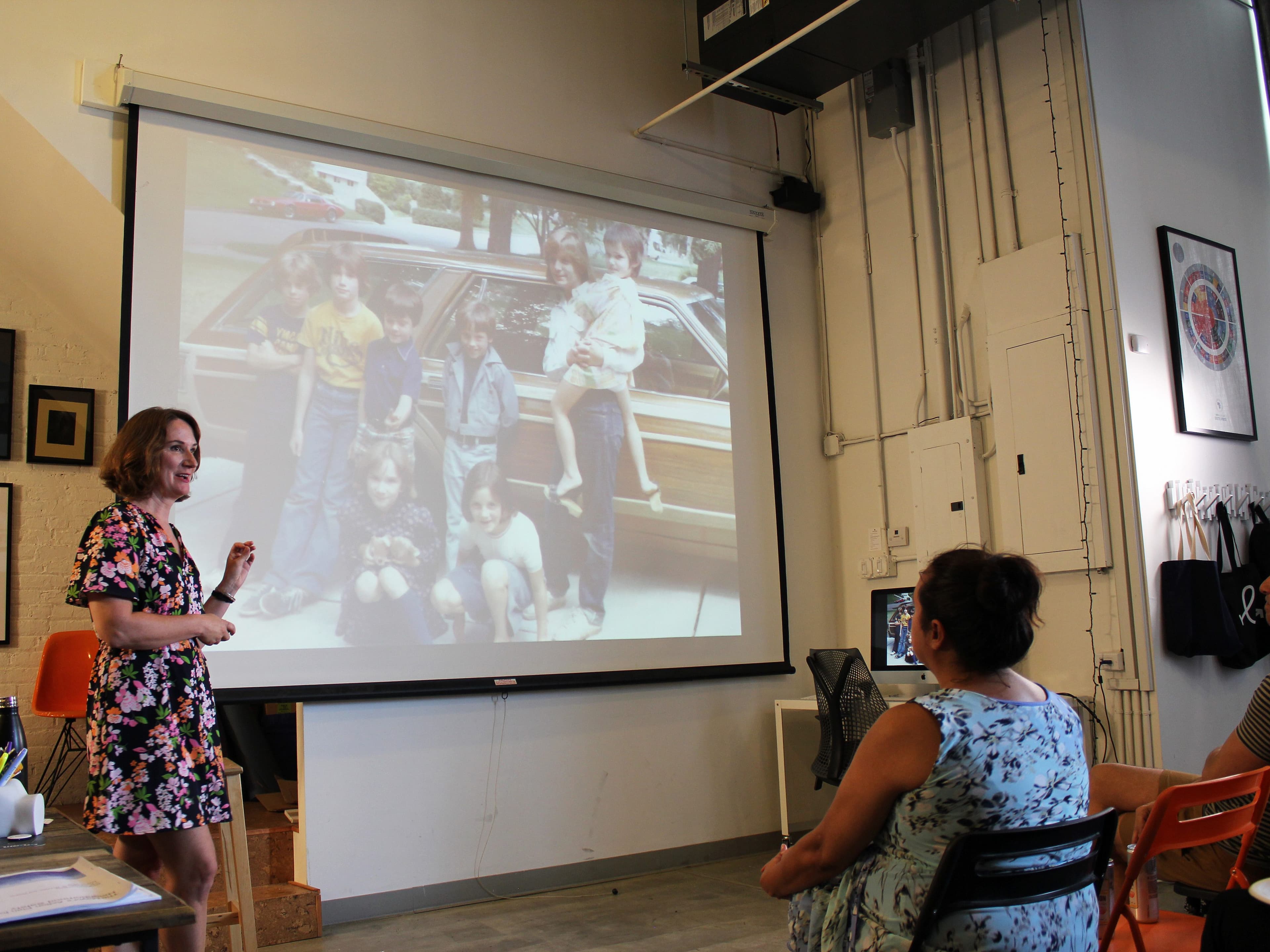 A woman in a floral dress speaks to a seated audience, pointing at a projected image of a family posing next to a station wagon. The setting appears to be a workshop or classroom with artwork on the walls and chairs arranged for attendees.