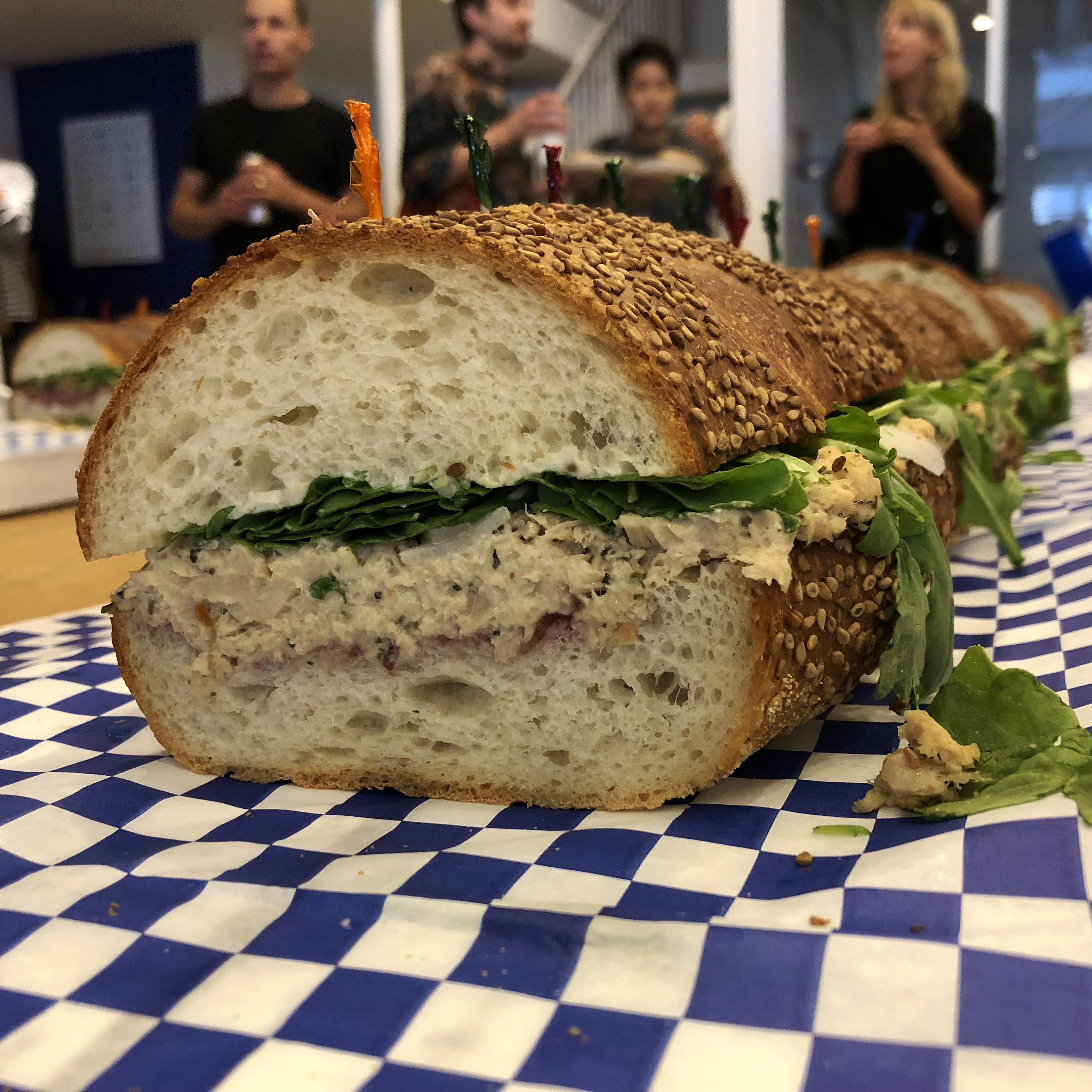 A close-up of a large sandwich cut into sections, with sesame seed-topped bread, filled with leafy greens and a creamy spread, placed on blue and white checkered paper. Several people are in the background, some talking and others taking photos.