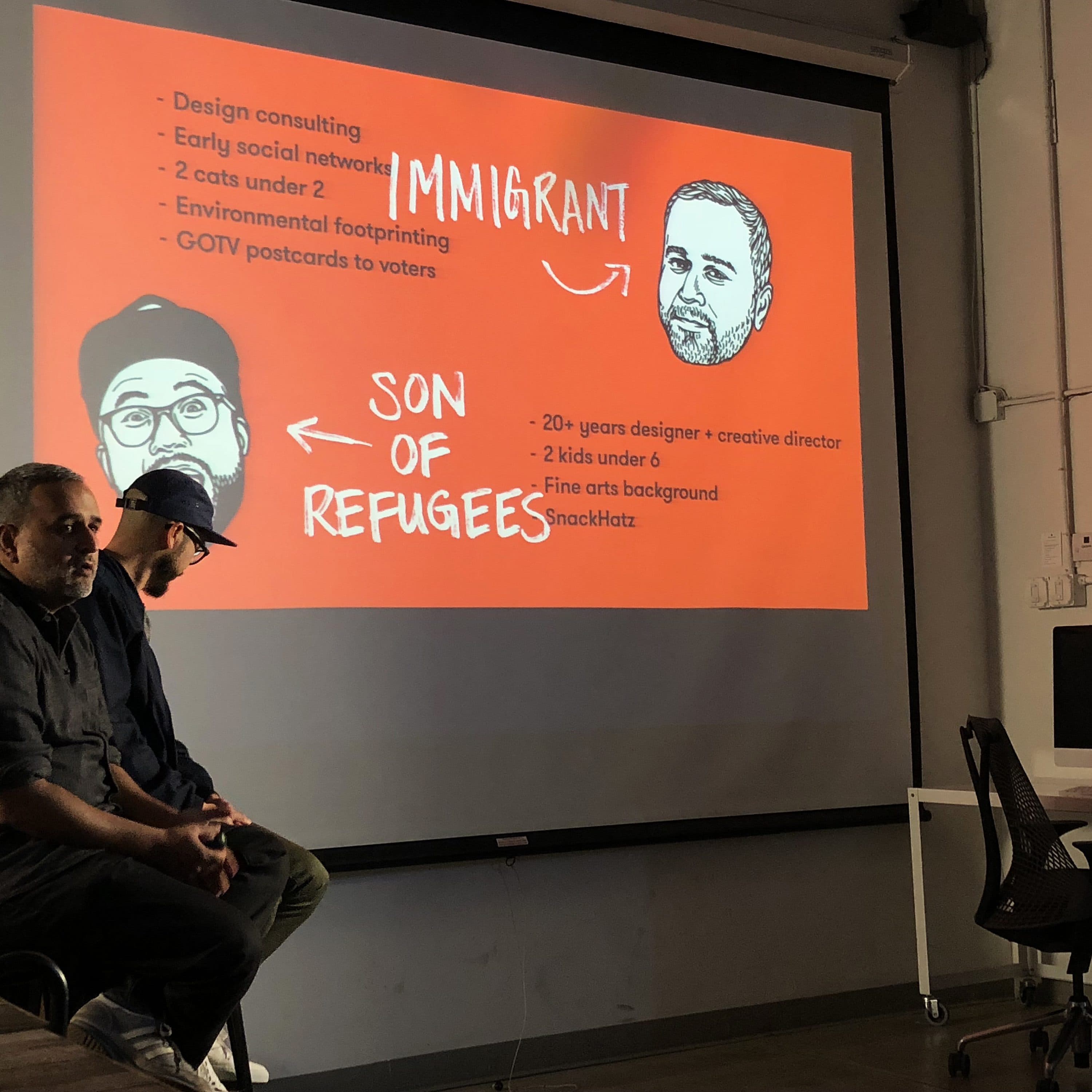 Two people are sitting in a classroom, giving a presentation. A projector screen displays cartoon-style graphics of two individuals with text. The text includes bullet points about design consulting, social networks, being an immigrant, and years of experience.